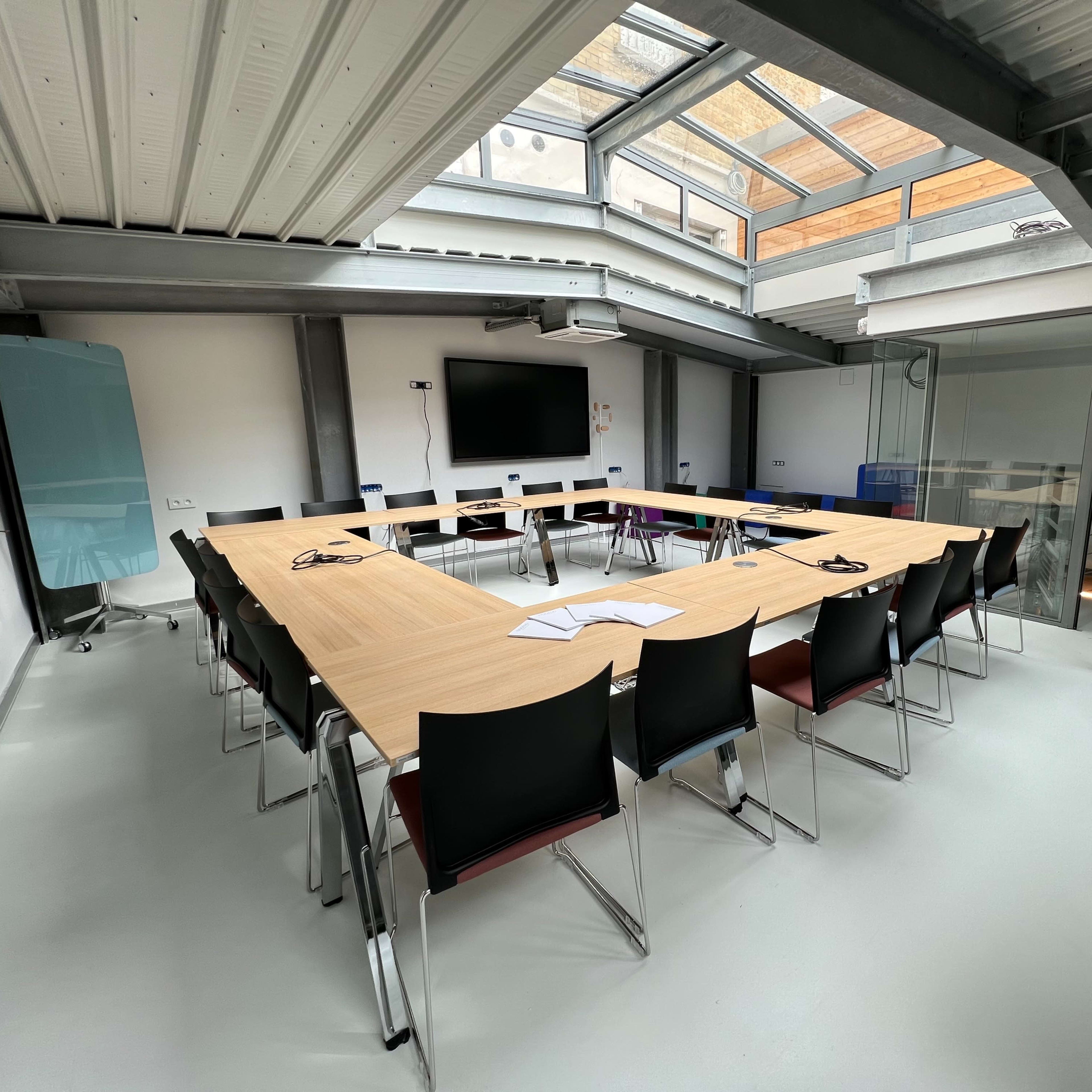 A modern conference room features a large rectangular table surrounded by black chairs, with a screen mounted on the wall and large windows above.