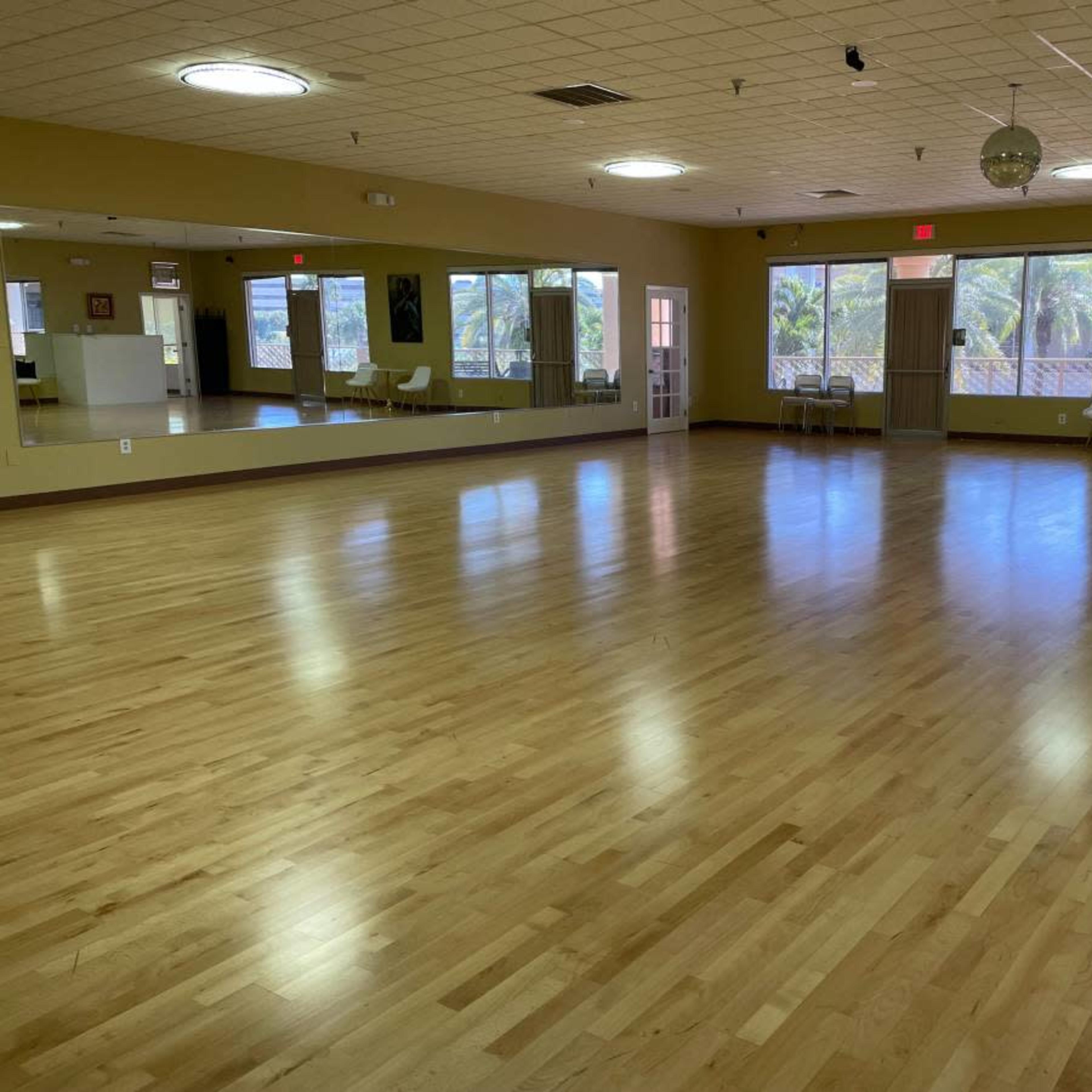 A spacious, empty dance studio with wooden flooring and large windows letting in natural light.