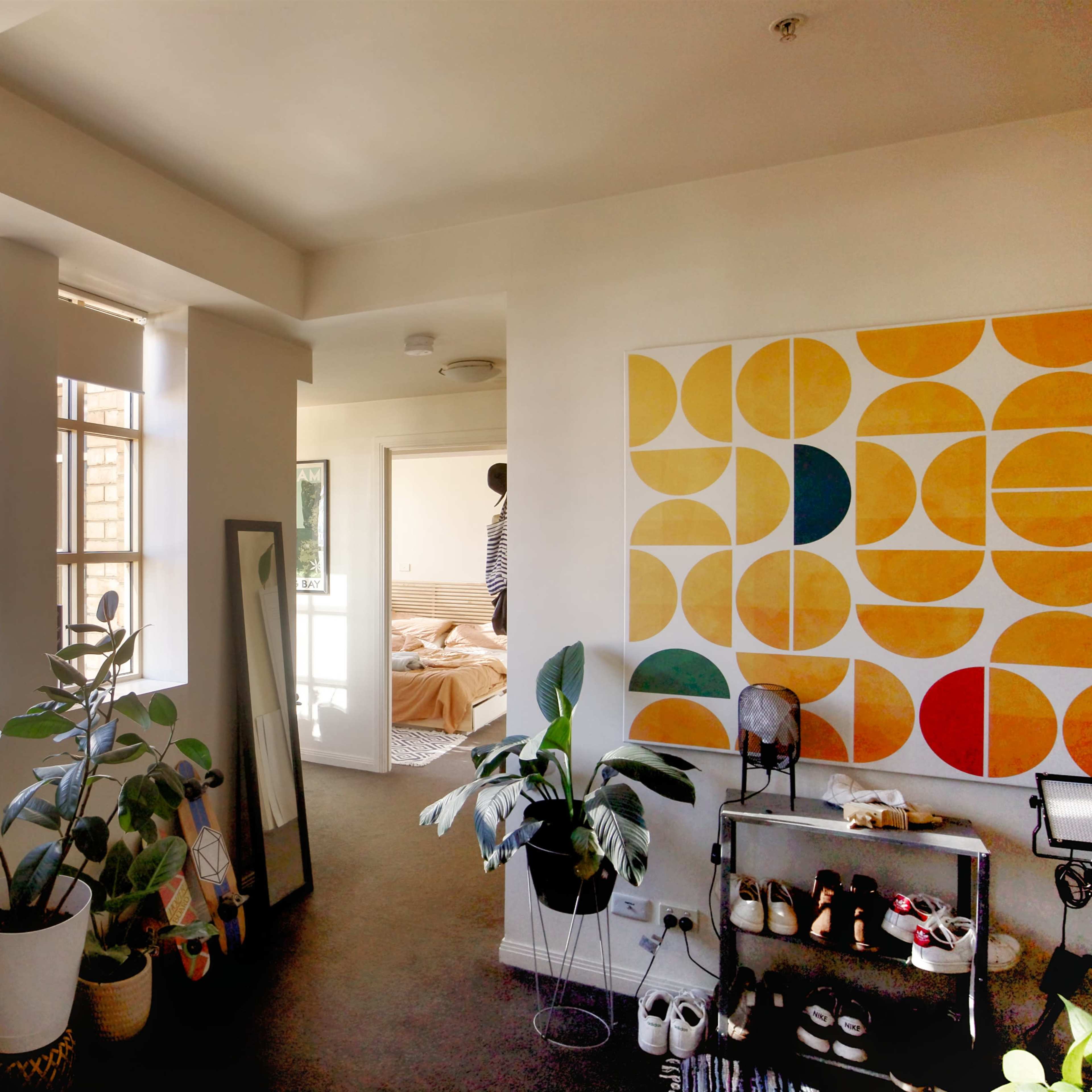 A bright interior of a room featuring large windows, plants, a mirror, a colorful geometric wall art, and a glimpse of a bedroom in the background.