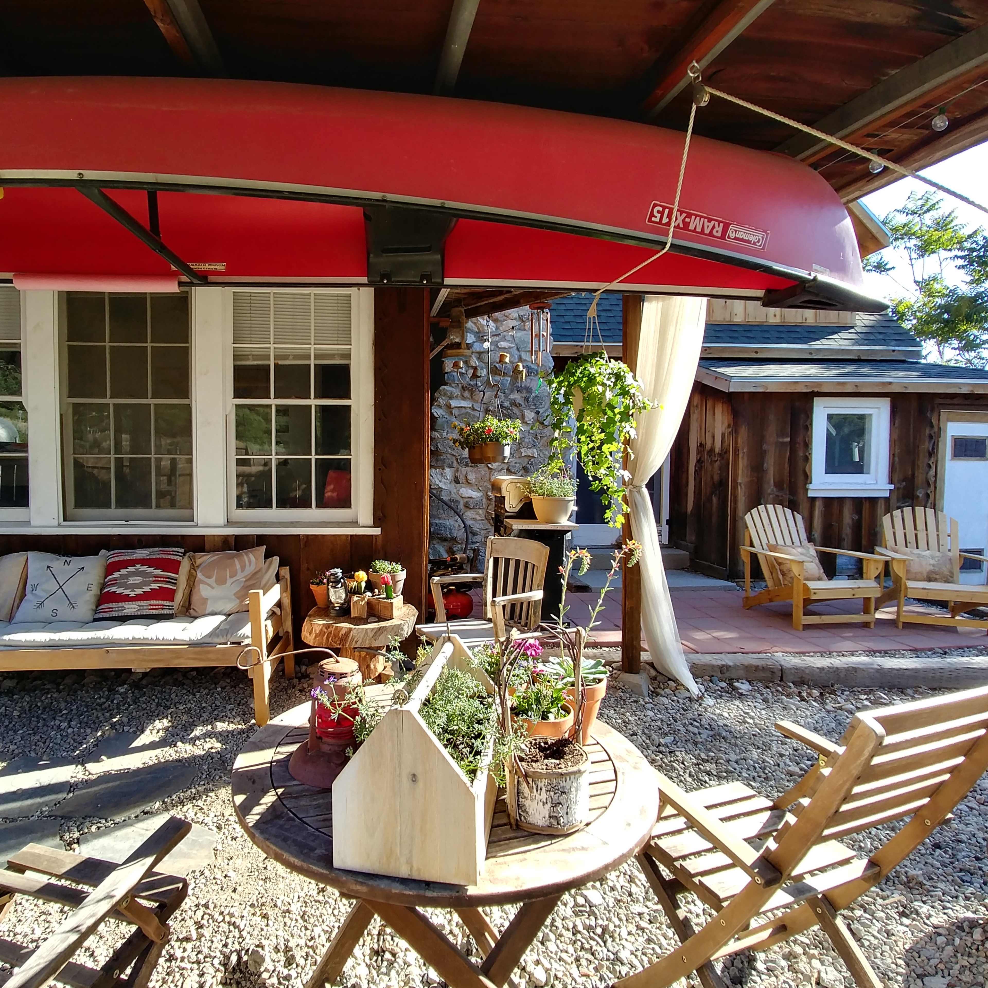 The scene features a cozy outdoor sitting area with a canoe suspended above a wooden bench, surrounded by potted plants and rustic seating.