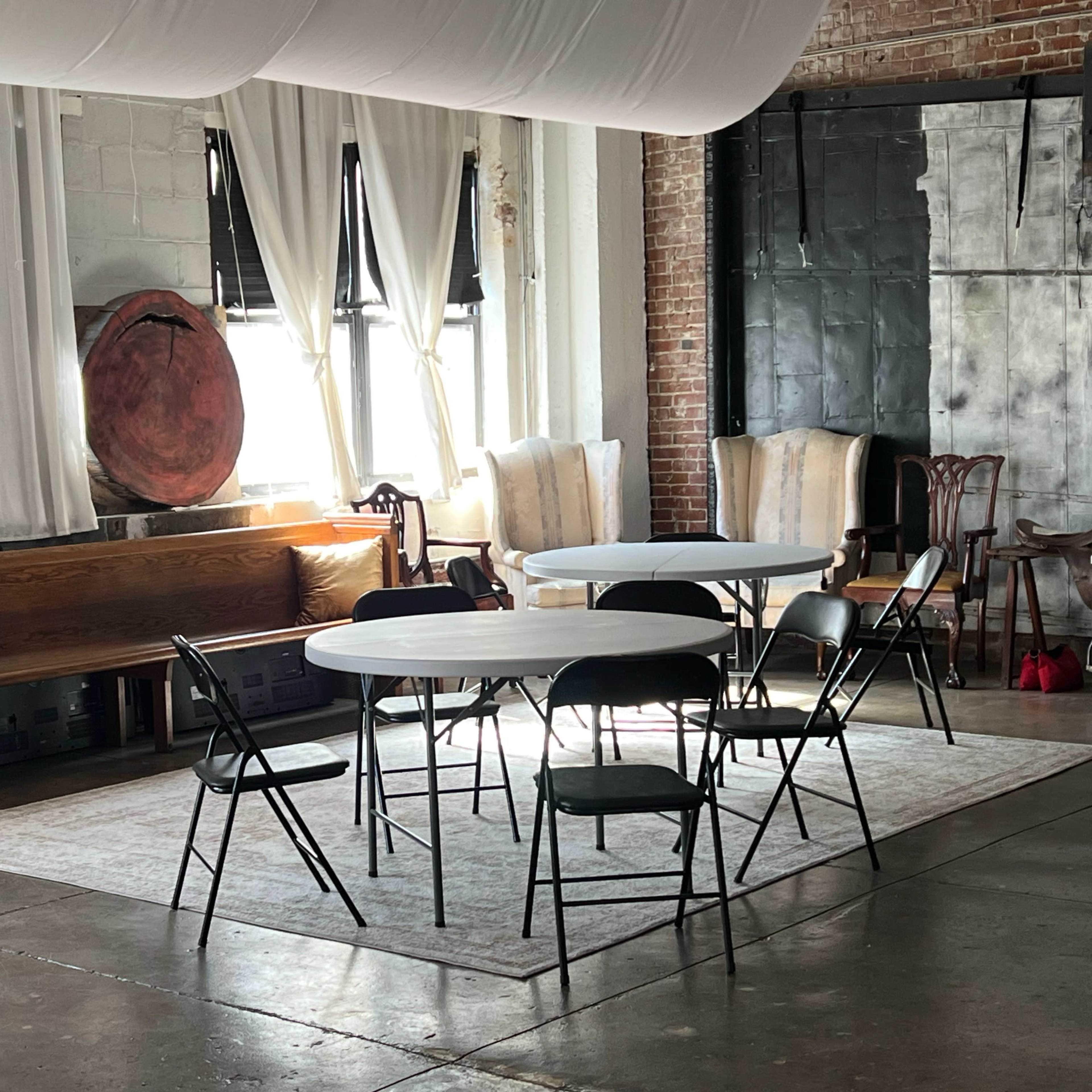 A spacious room features two round tables surrounded by black folding chairs on a light-colored rug, with large windows allowing natural light to enter.