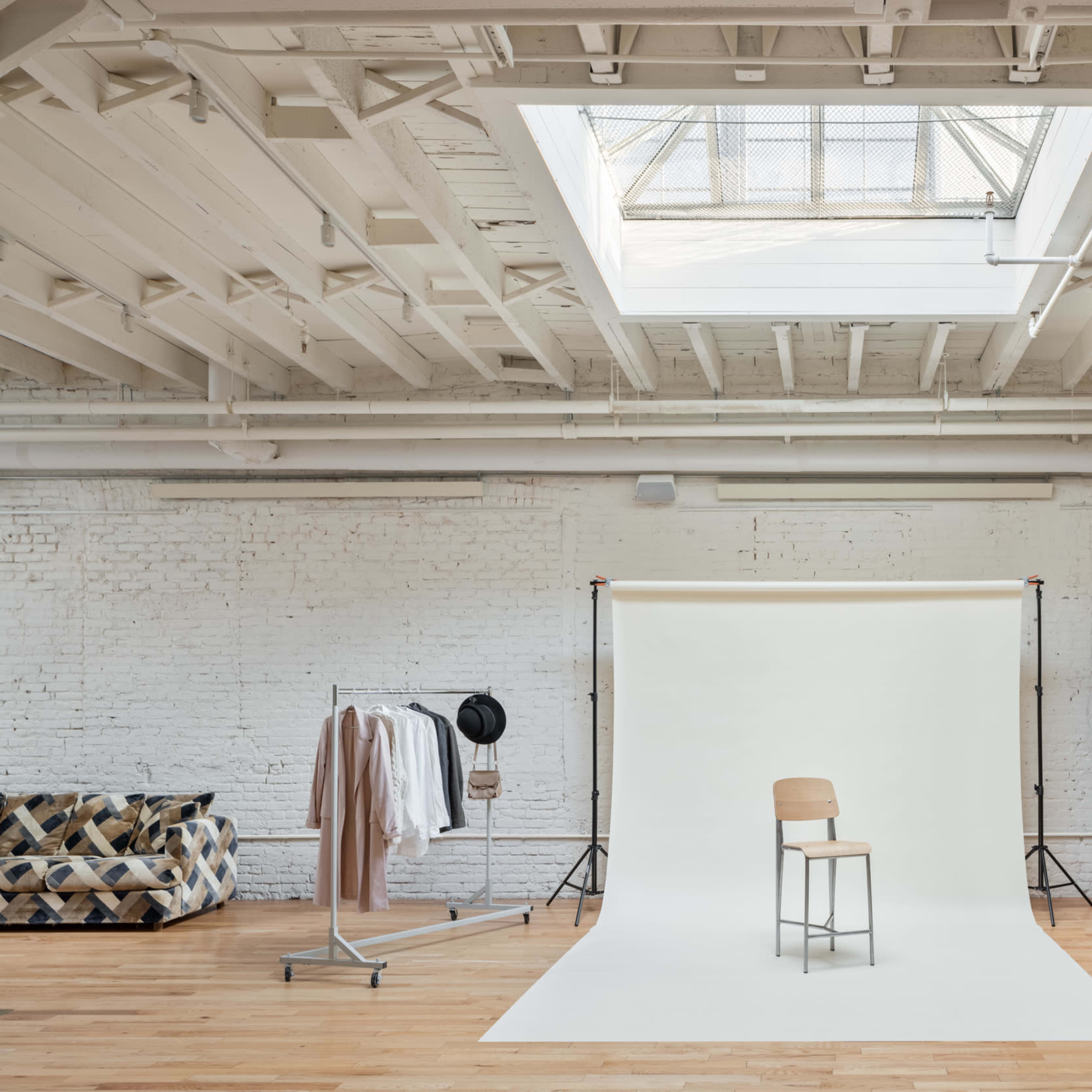 A photography studio featuring a wooden chair on a white backdrop, surrounded by clothing on a rack, a large light source, and a potted plant.