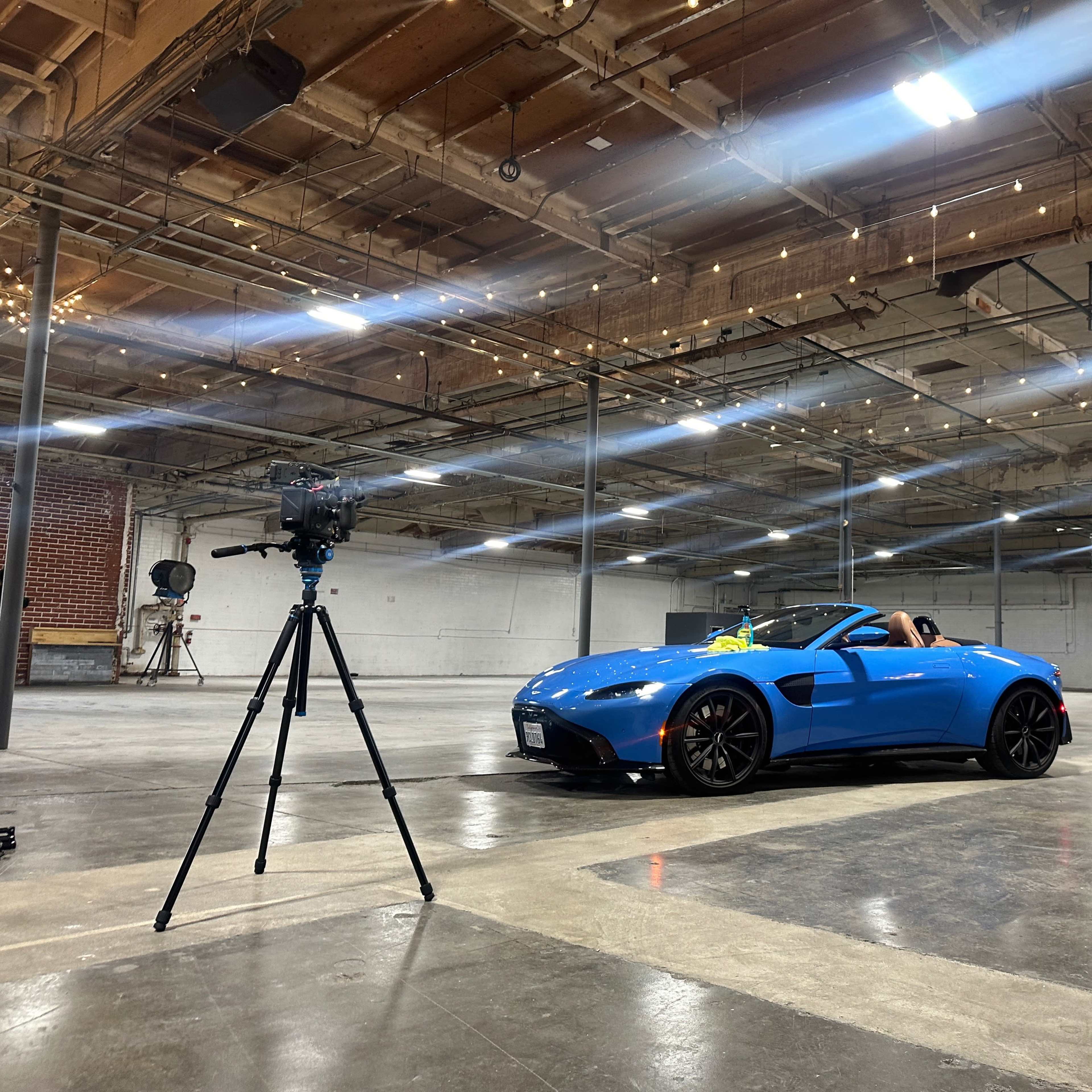 A blue convertible sports car is parked in an empty industrial space with a tripod-mounted camera positioned nearby.