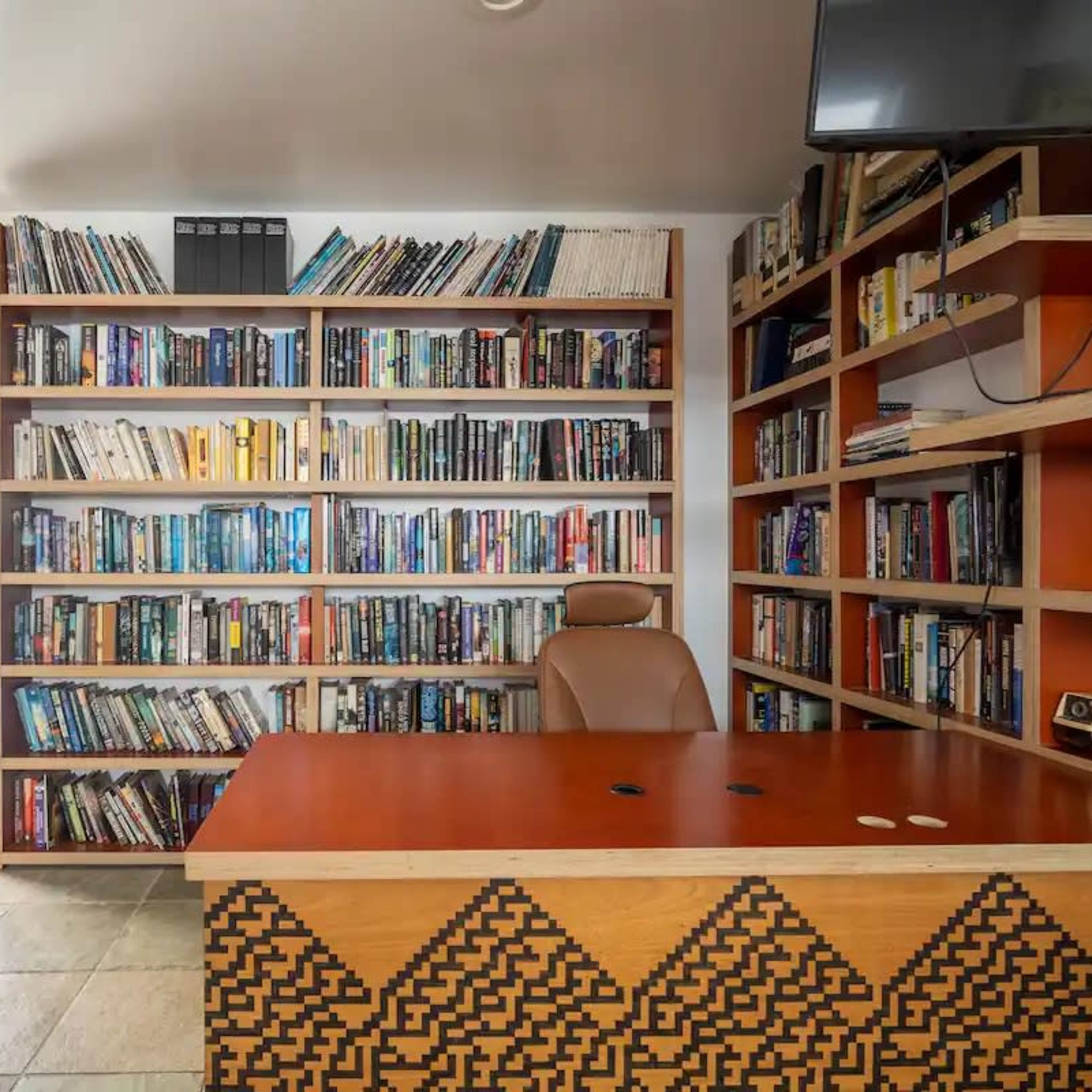A modern office with a wooden desk, an ergonomic chair, and a wall-mounted television, surrounded by shelves filled with books.