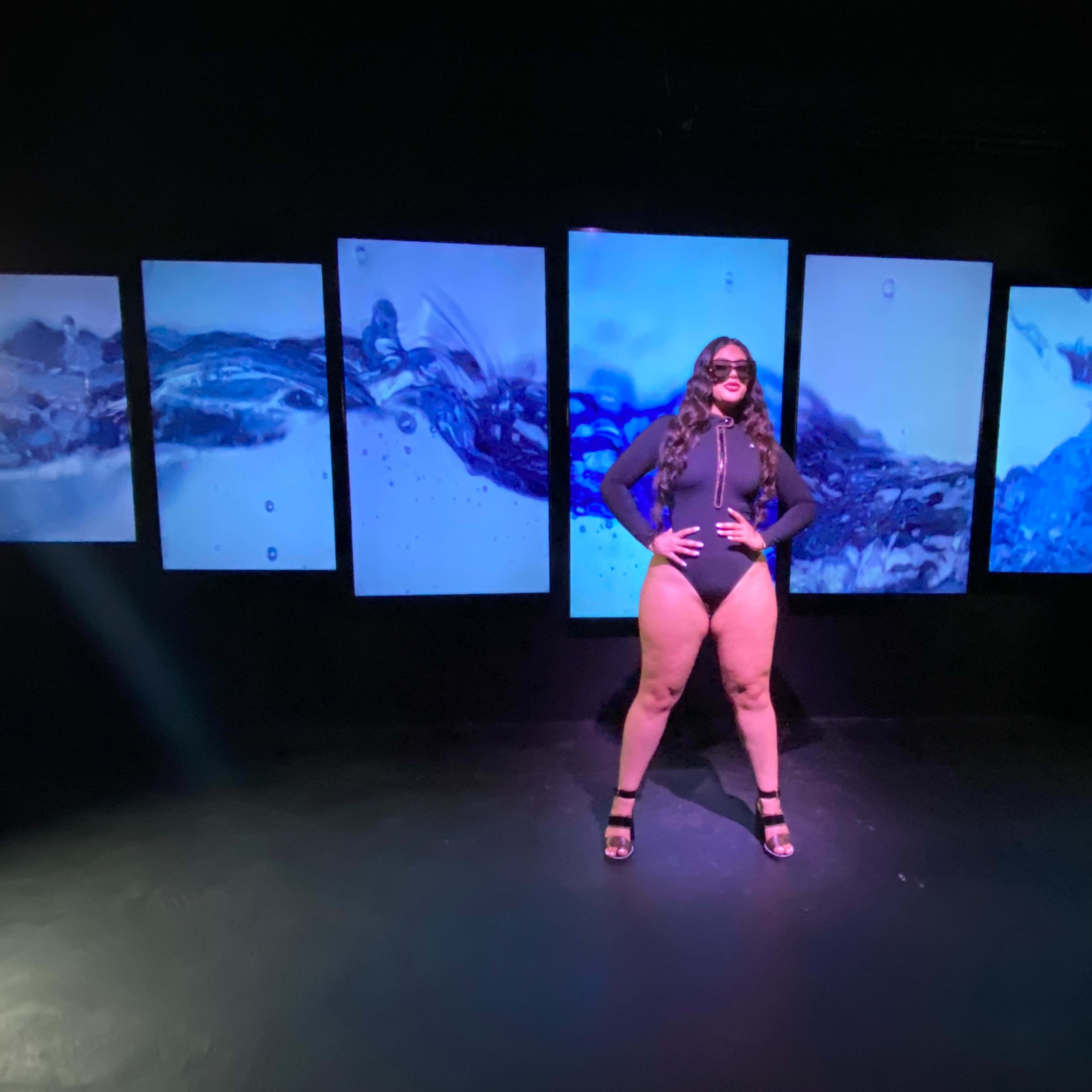 A person in a black swimsuit poses in front of a series of screens displaying blue water-themed imagery.