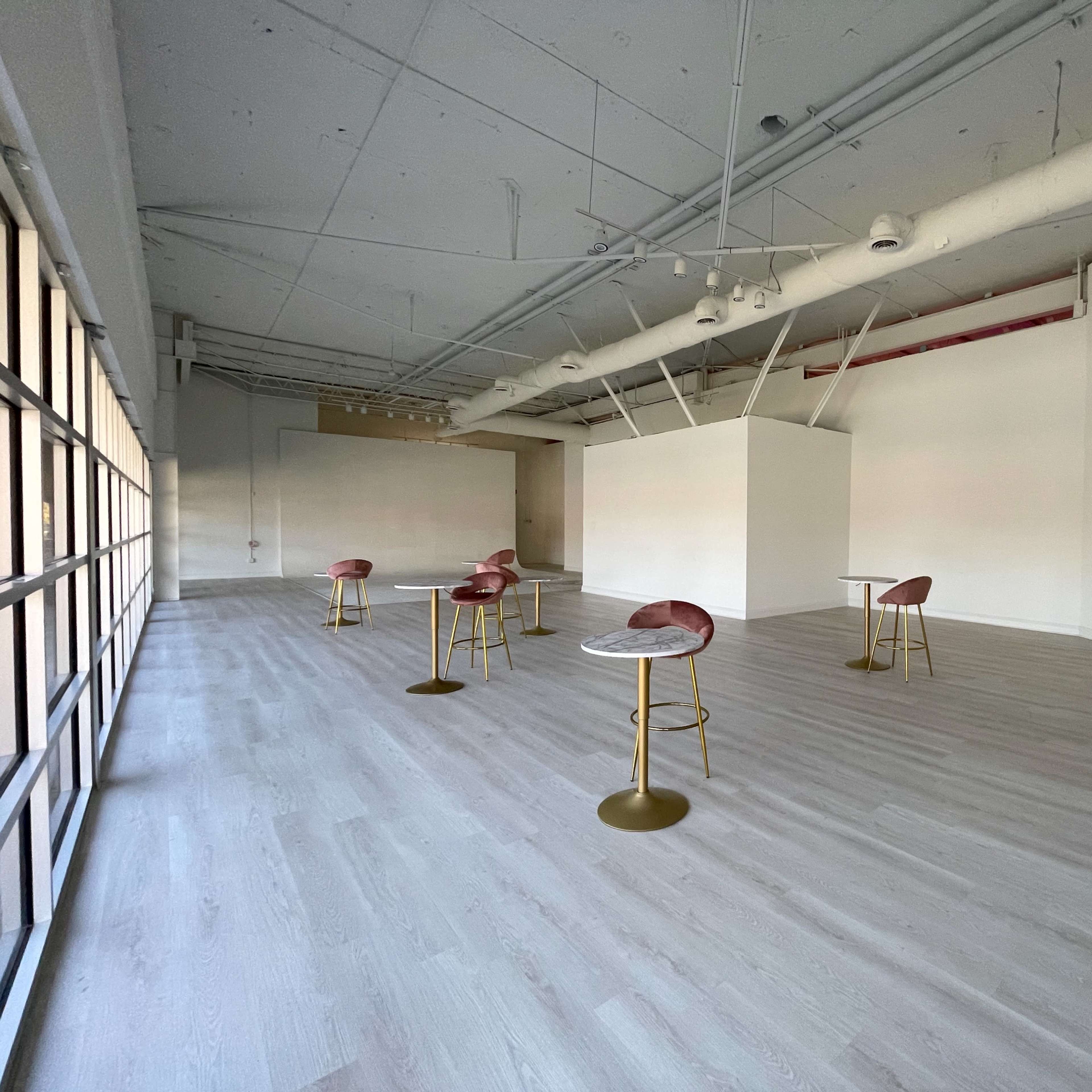 An empty modern room features several tall small tables with pink seating arranged on a light-colored wooden floor.