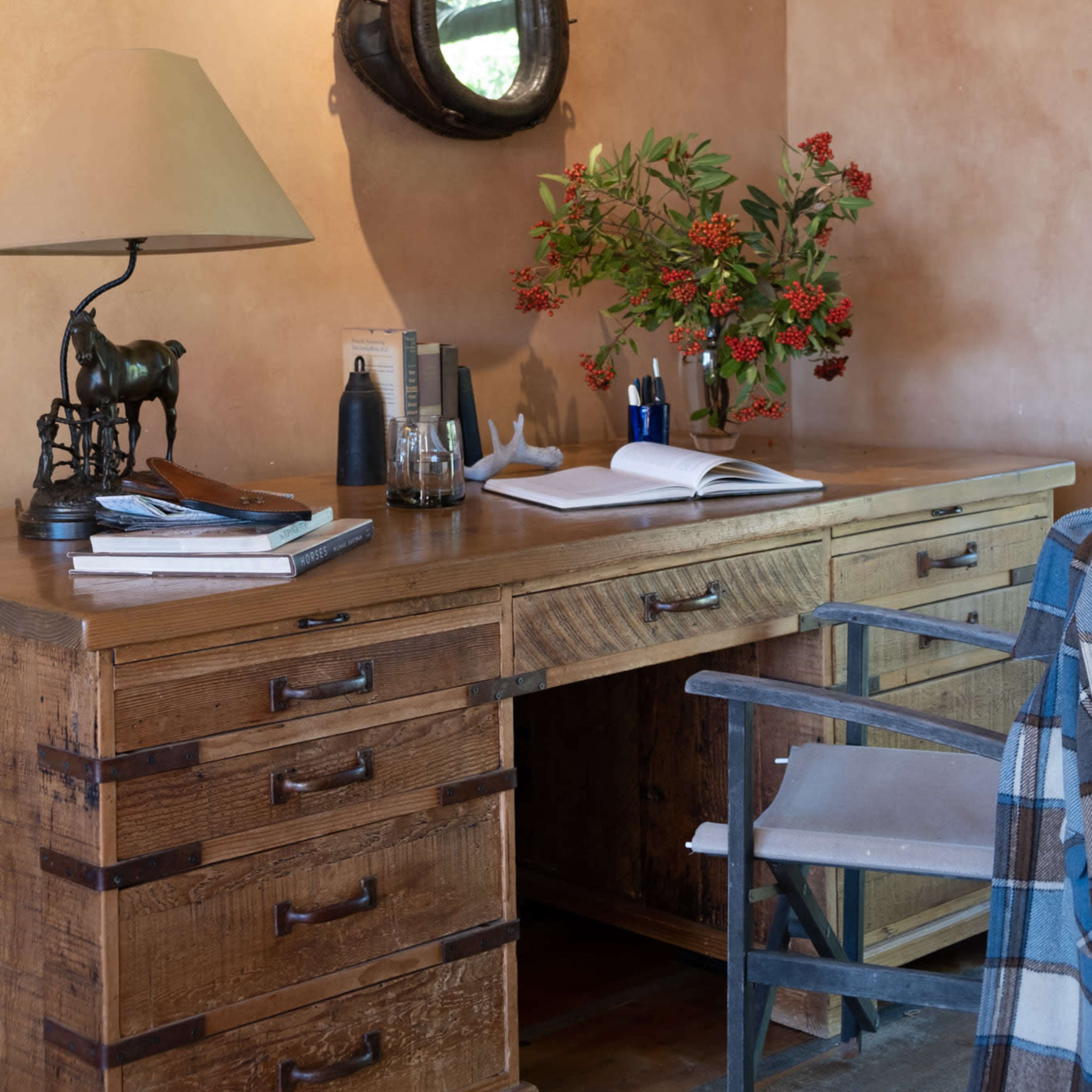 A wooden desk with drawers is adorned with a lamp, a small horse sculpture, a vase of red flowers, and an open notebook, while a wall-mounted mirror hangs above.