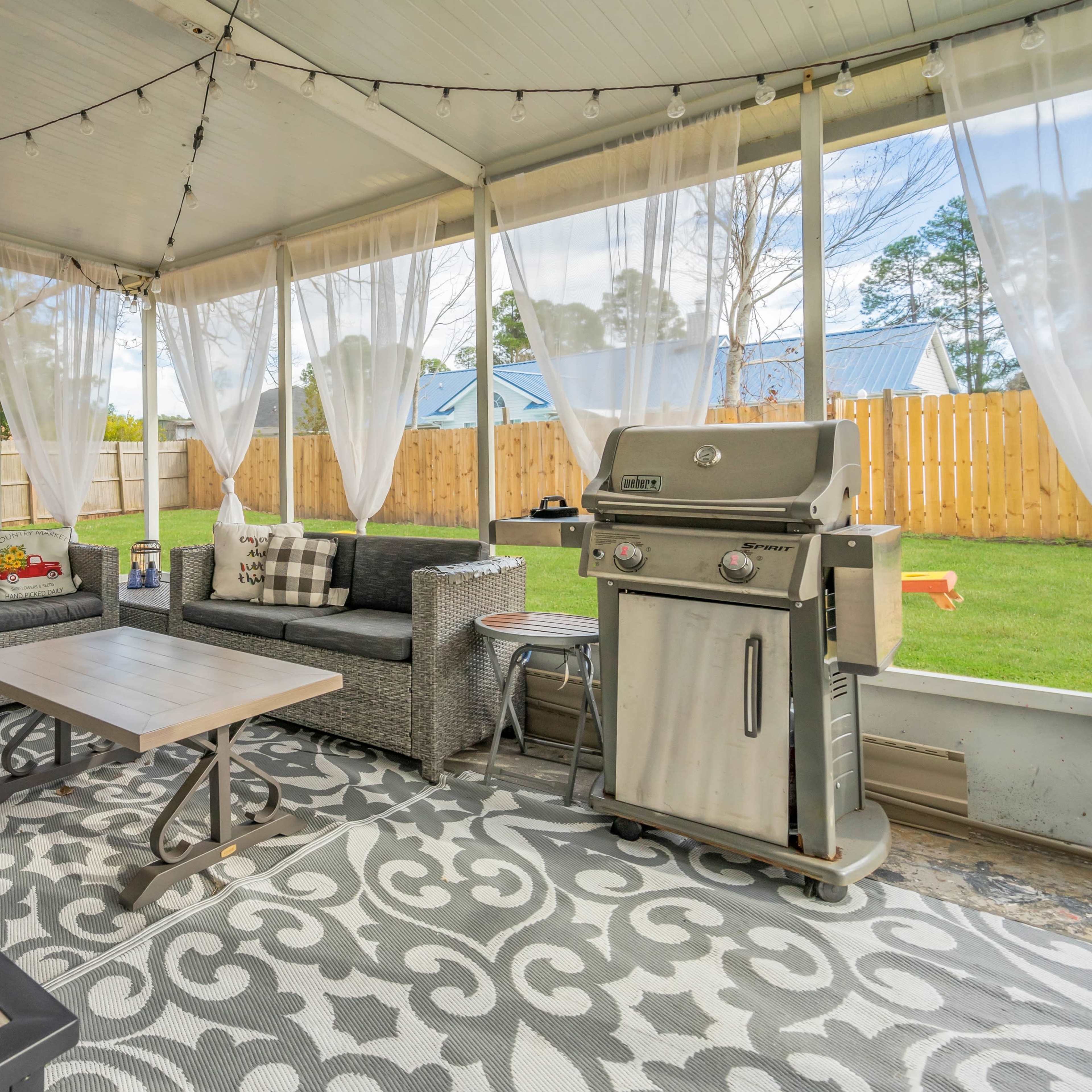 A covered outdoor patio featuring a grill, seating area with gray couches, a coffee table, and a decorative rug, all surrounded by a backyard with a wooden fence.