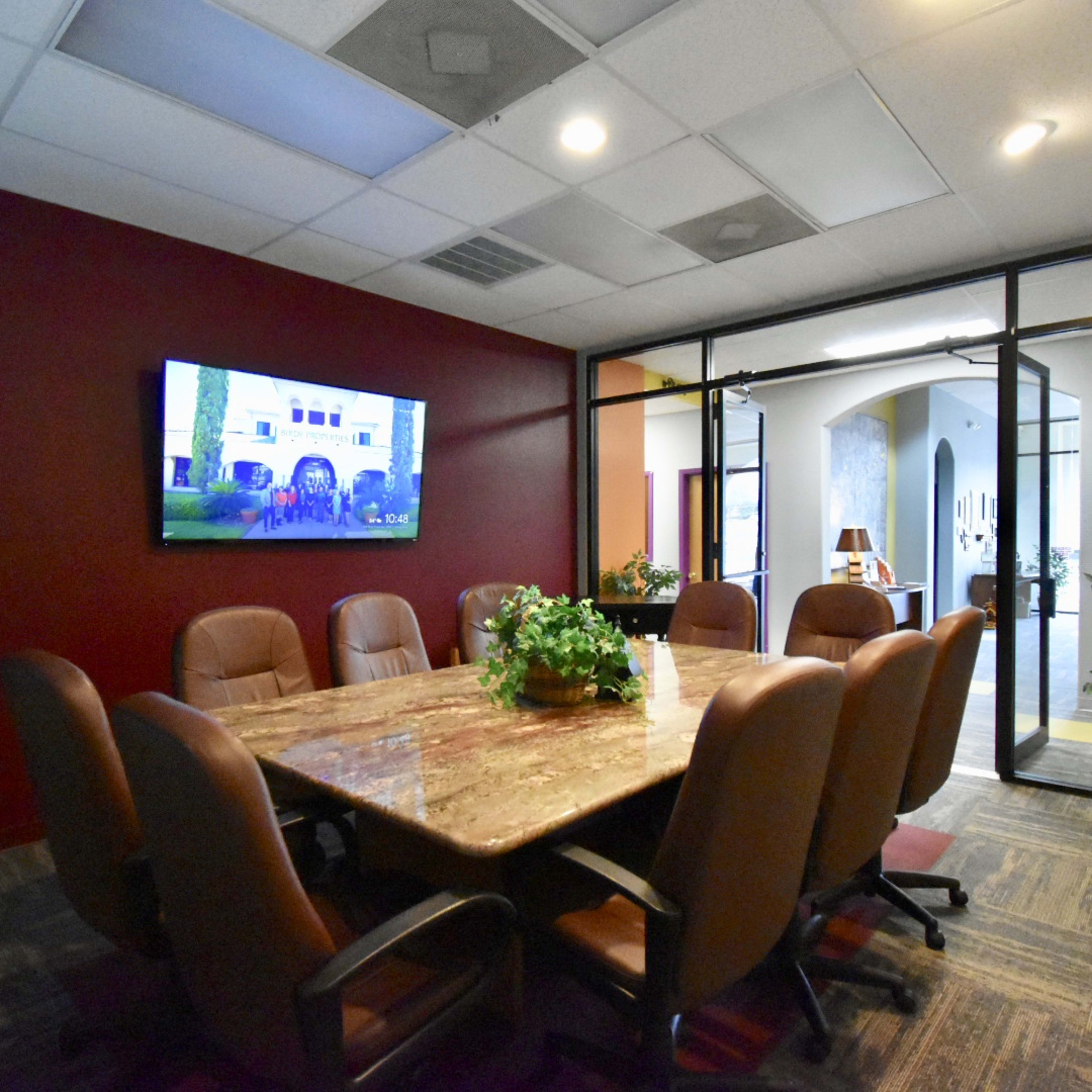 A conference room features a large table surrounded by leather chairs and a wall-mounted TV in a space with dark red walls.