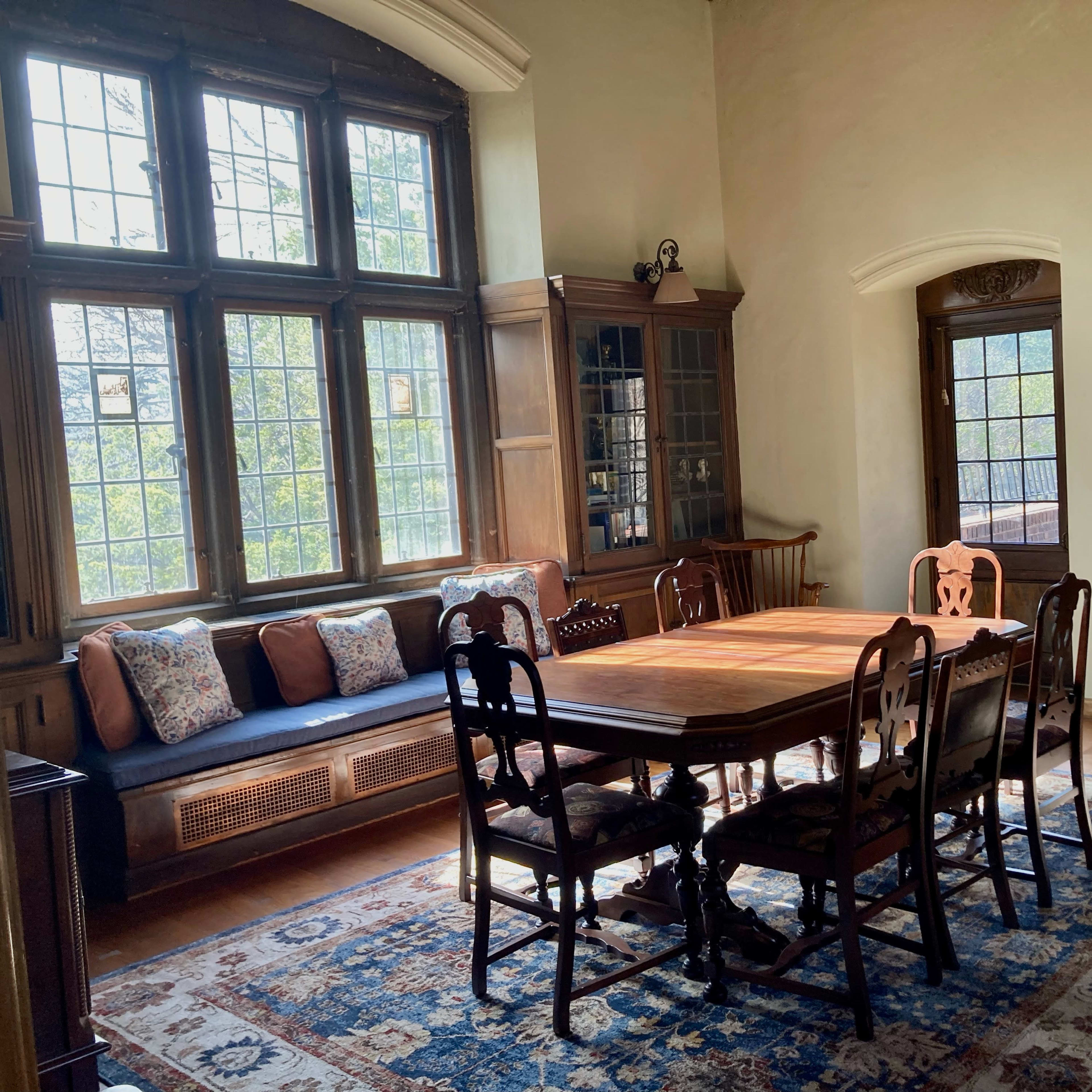 A spacious room features a wooden dining table surrounded by six chairs, with a window seat and large windows letting in natural light.