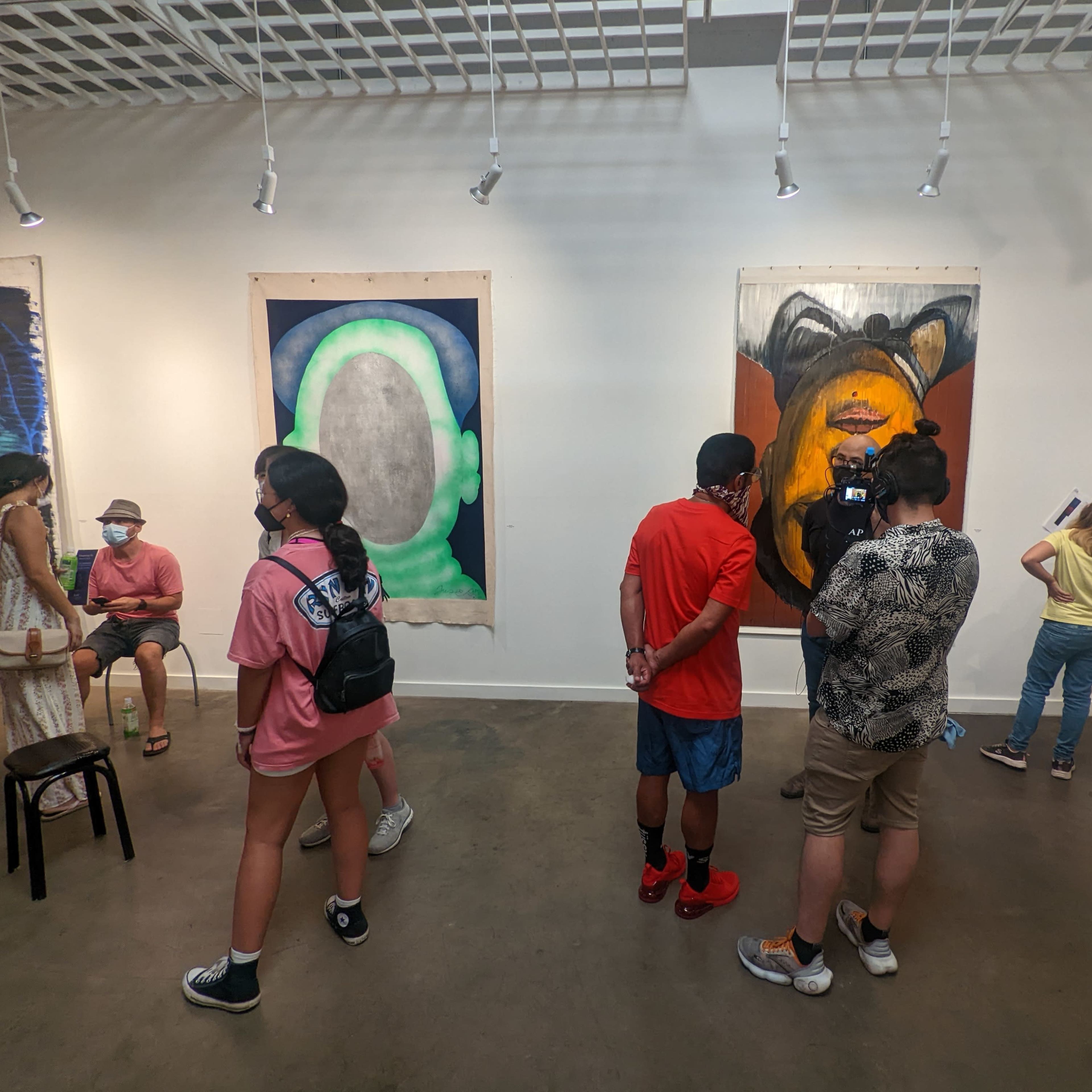 An art gallery featuring several people viewing large paintings on the walls, while others are sitting or engaged in conversation.
