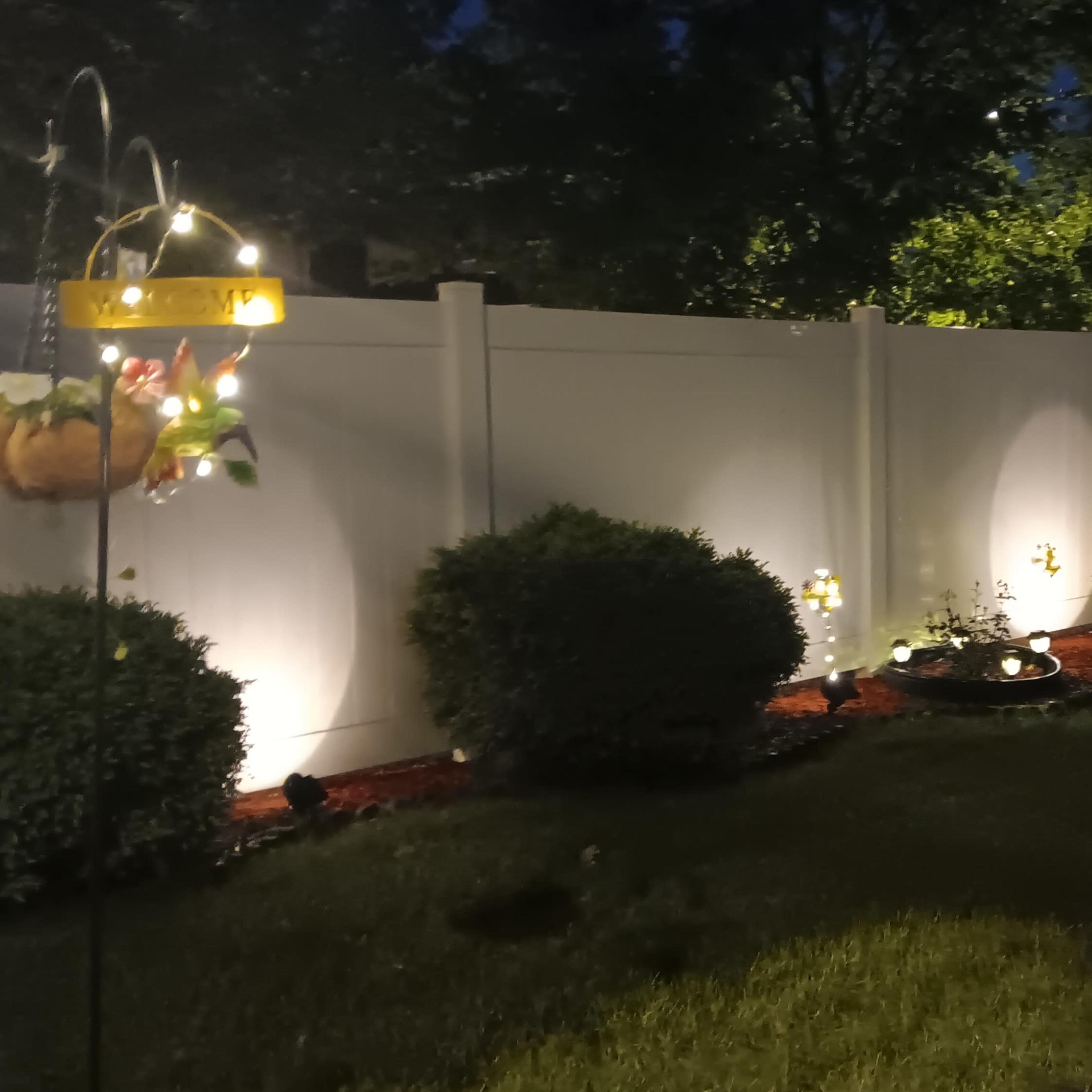 A well-lit garden area at night, featuring a white fence, neatly trimmed bushes, and decorative light fixtures.