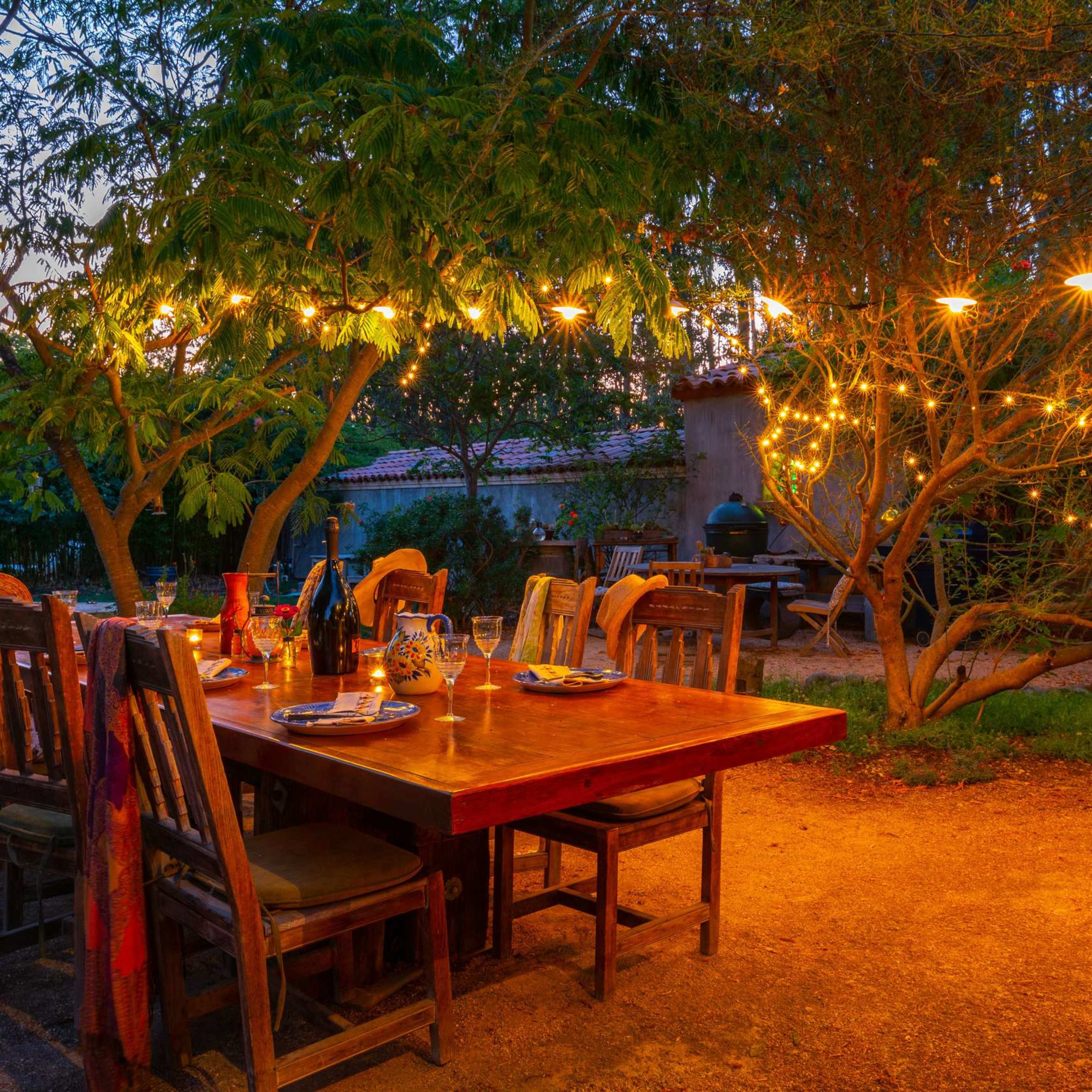 A wooden dining table is set outdoors under string lights, surrounded by chairs and a tree, in a warm, twilight setting.