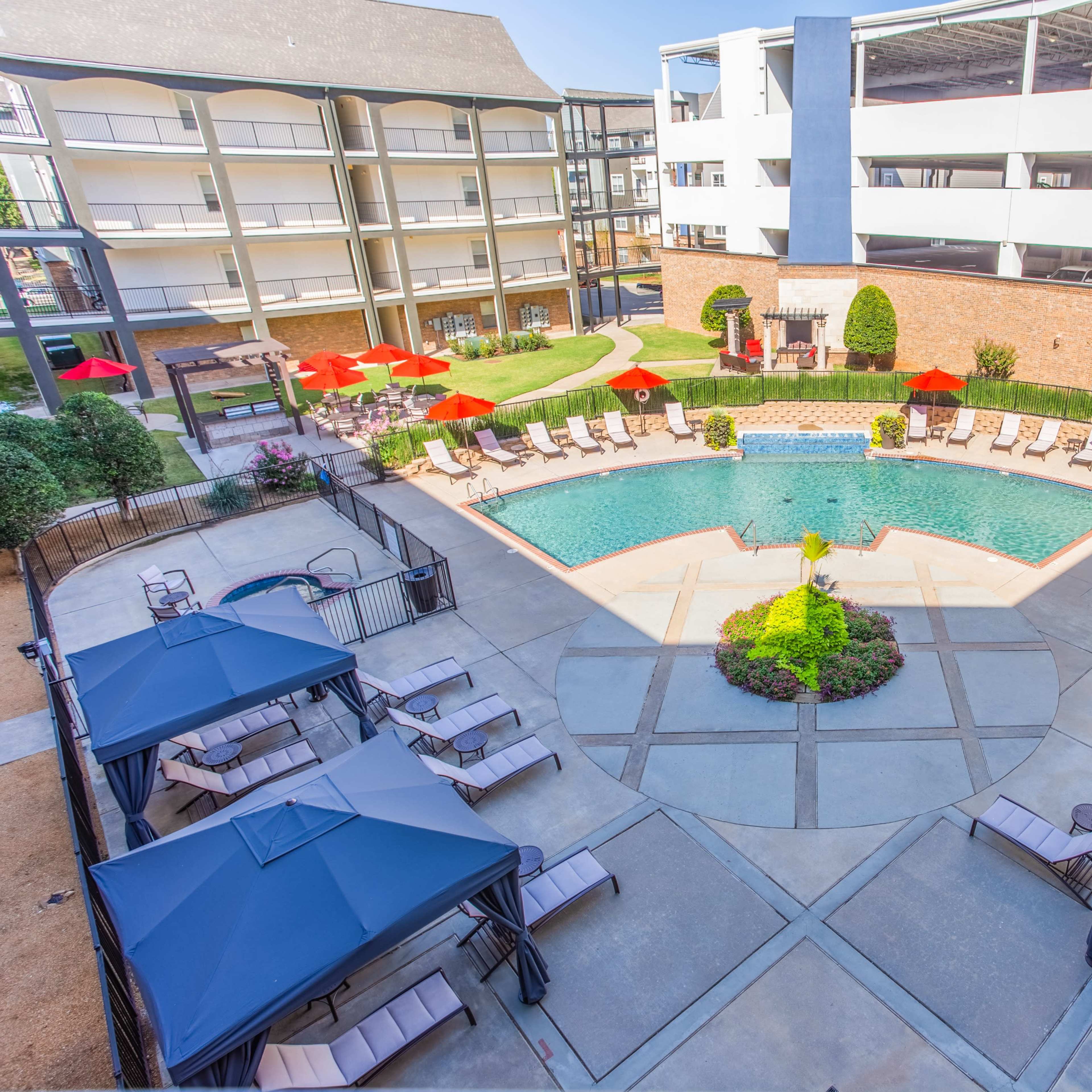An outdoor pool area surrounded by lounge chairs and umbrella canopies, with red umbrellas providing shade and a parking structure visible in the background.