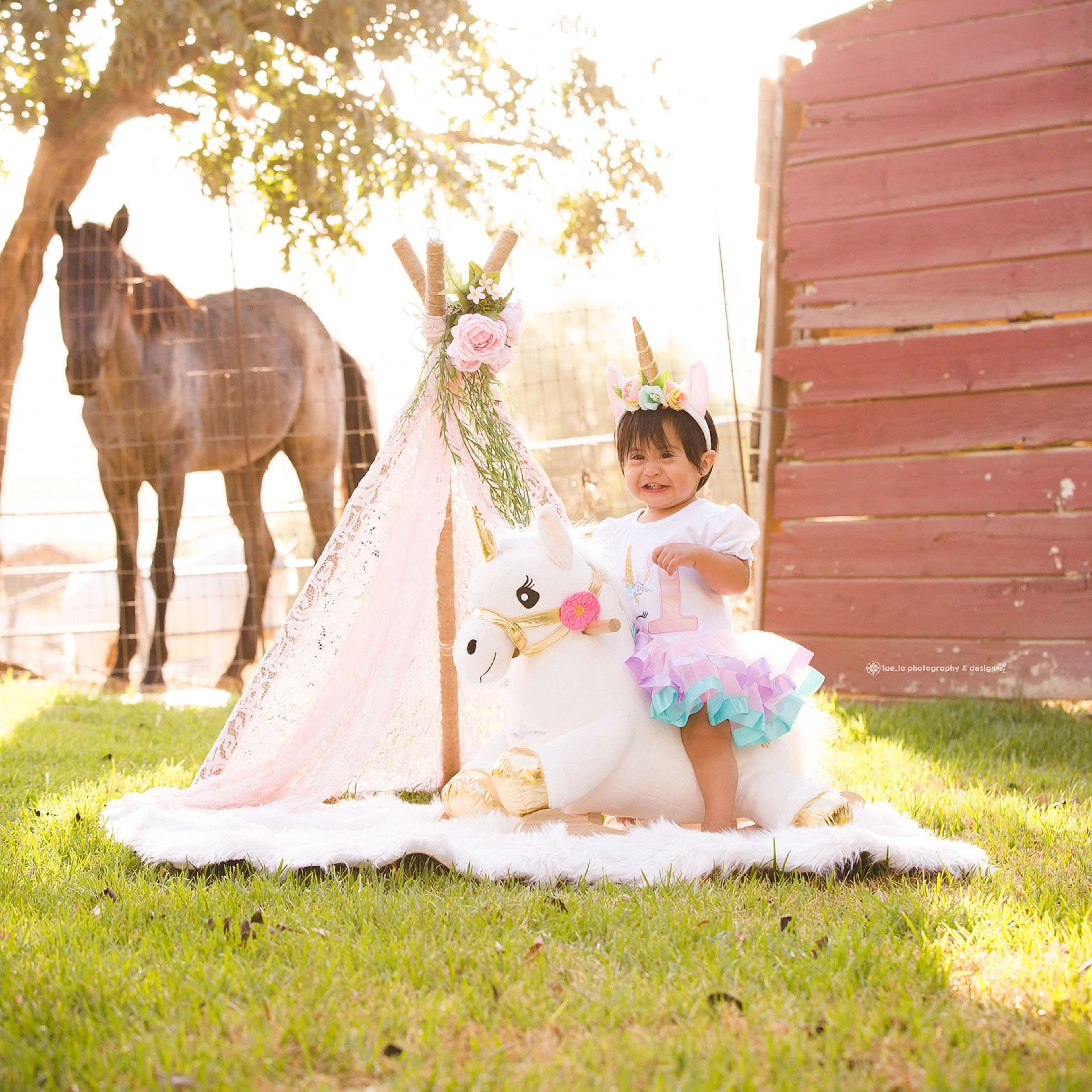 A young child stands on a rug in front of a unicorn-themed setup, with a horse in the background and sunlight filtering through the trees.