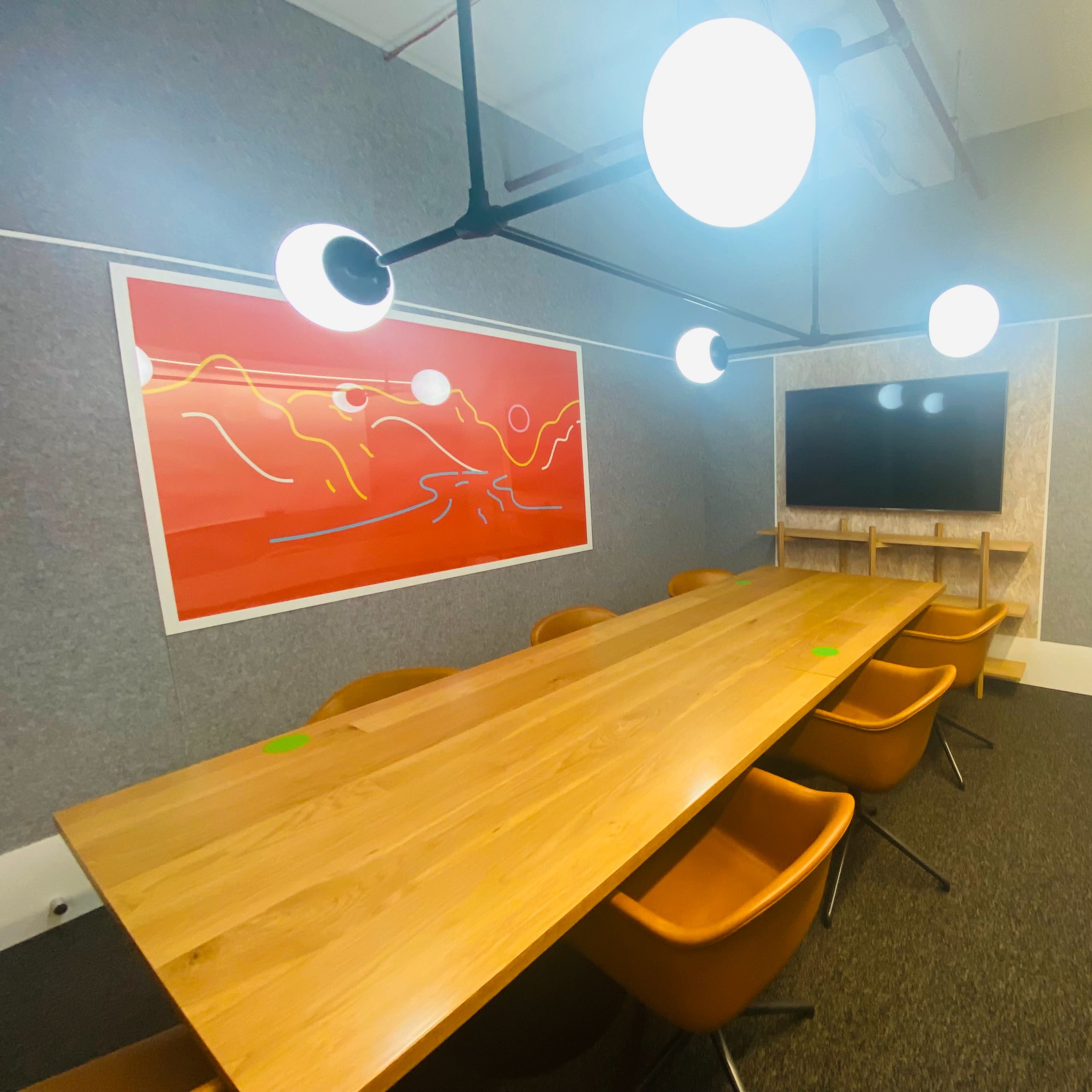 A modern conference room features a long wooden table surrounded by orange chairs, with a large screen mounted on the wall and bright overhead lighting.