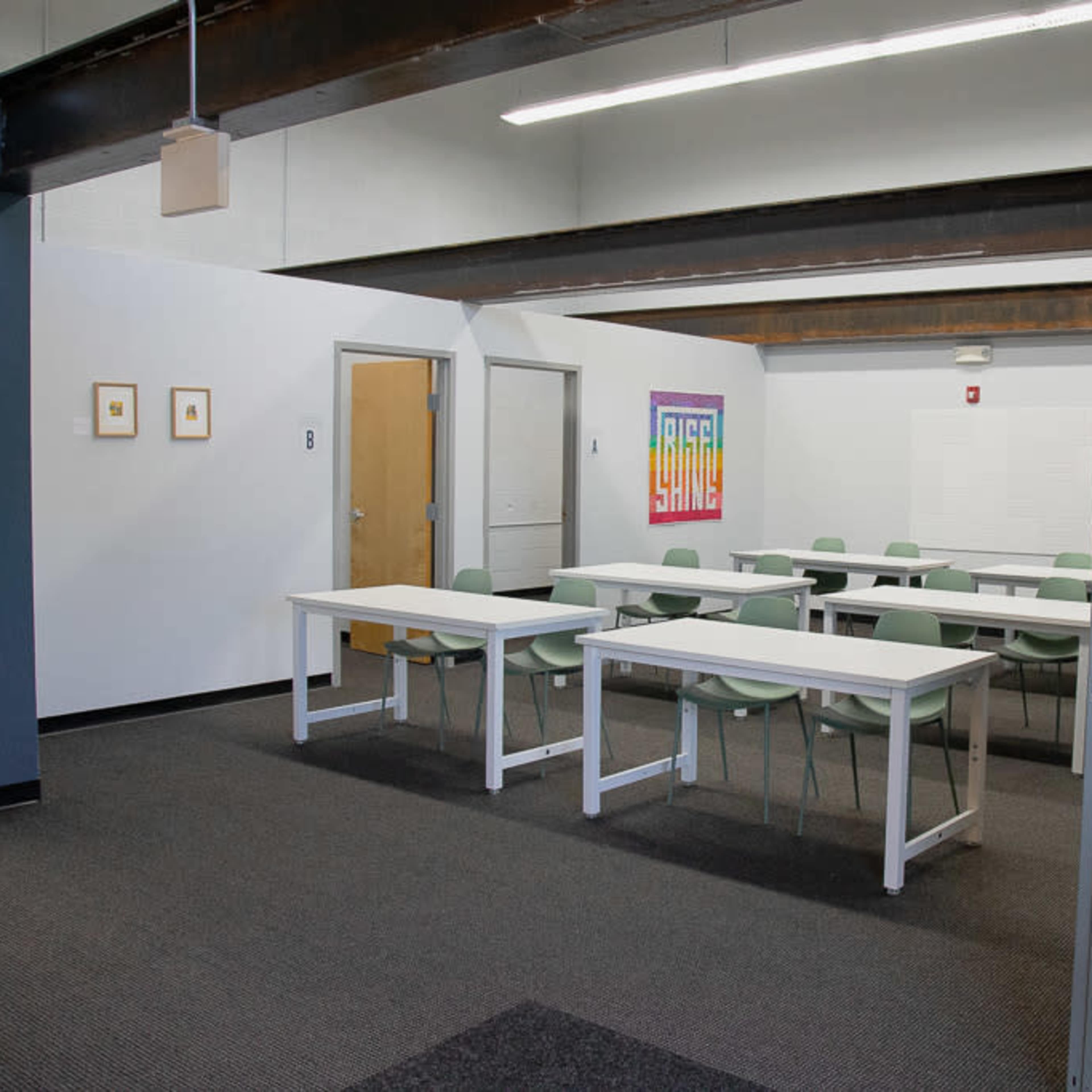 A classroom with several white tables and green chairs arranged in neat rows, and a colorful artwork displayed on the wall.
