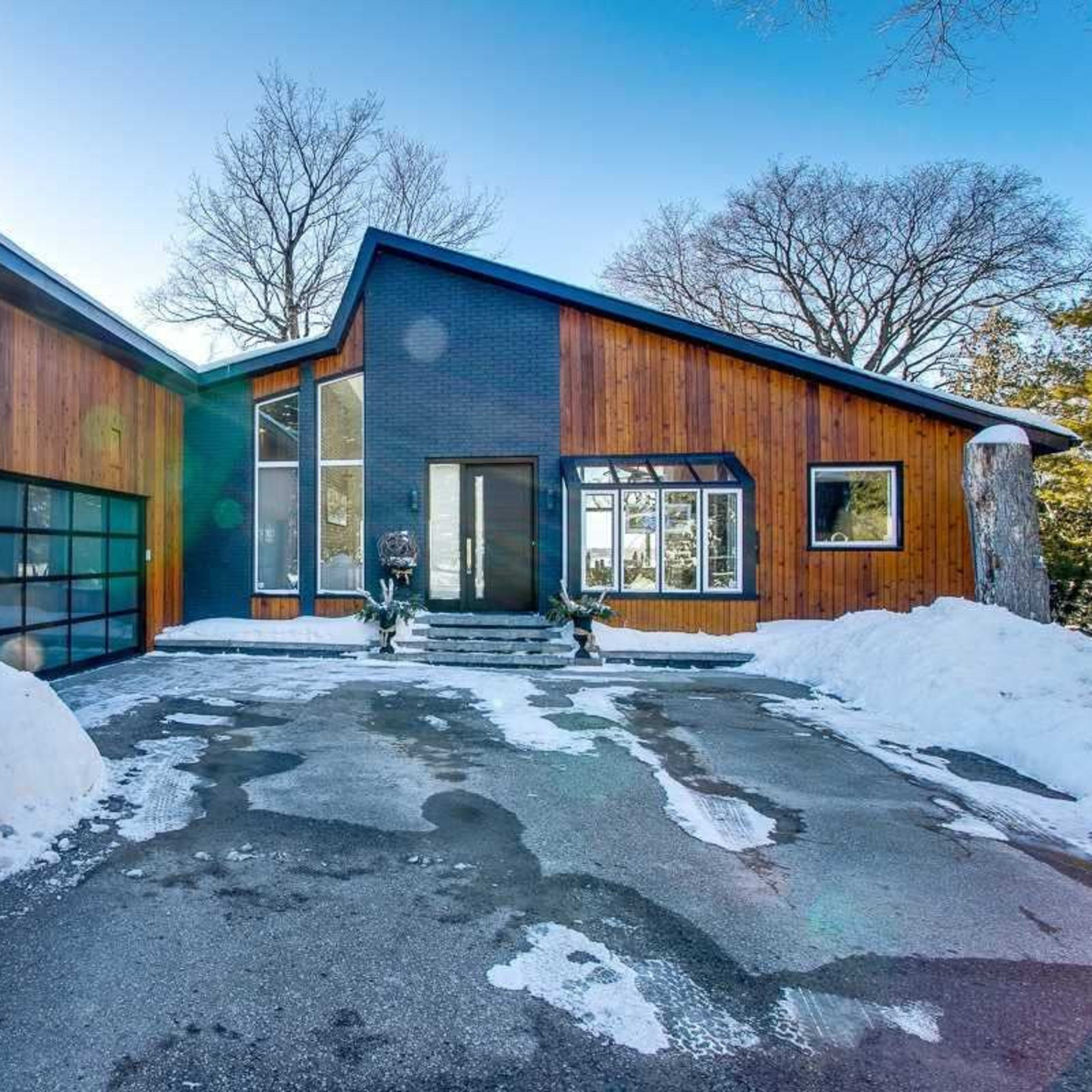 A modern house with wooden siding and a large garage sits on a snowy driveway under clear blue skies.