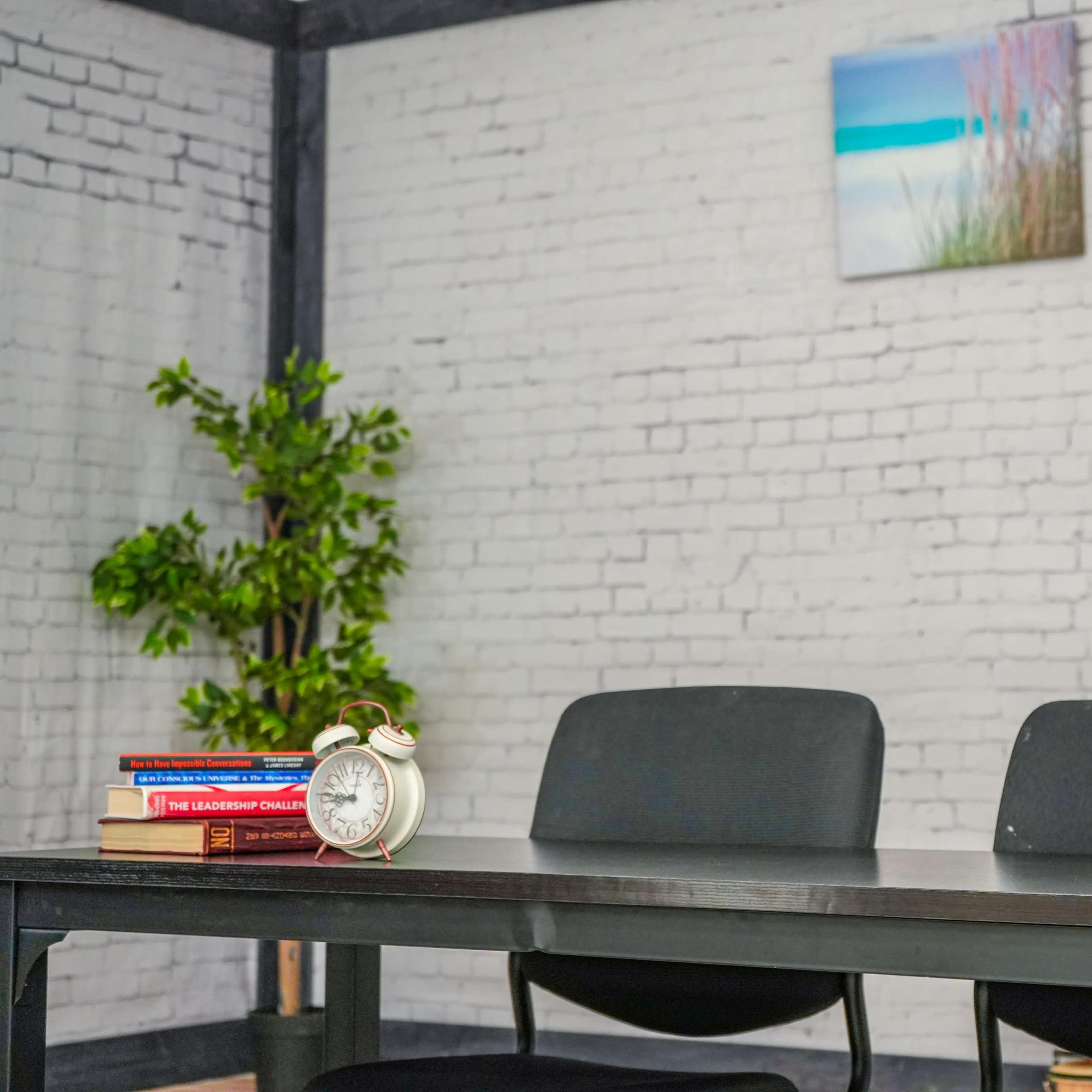 A minimalist office space features a black table, two chairs, a potted plant, an alarm clock, and a wall with a painting and exposed brick.