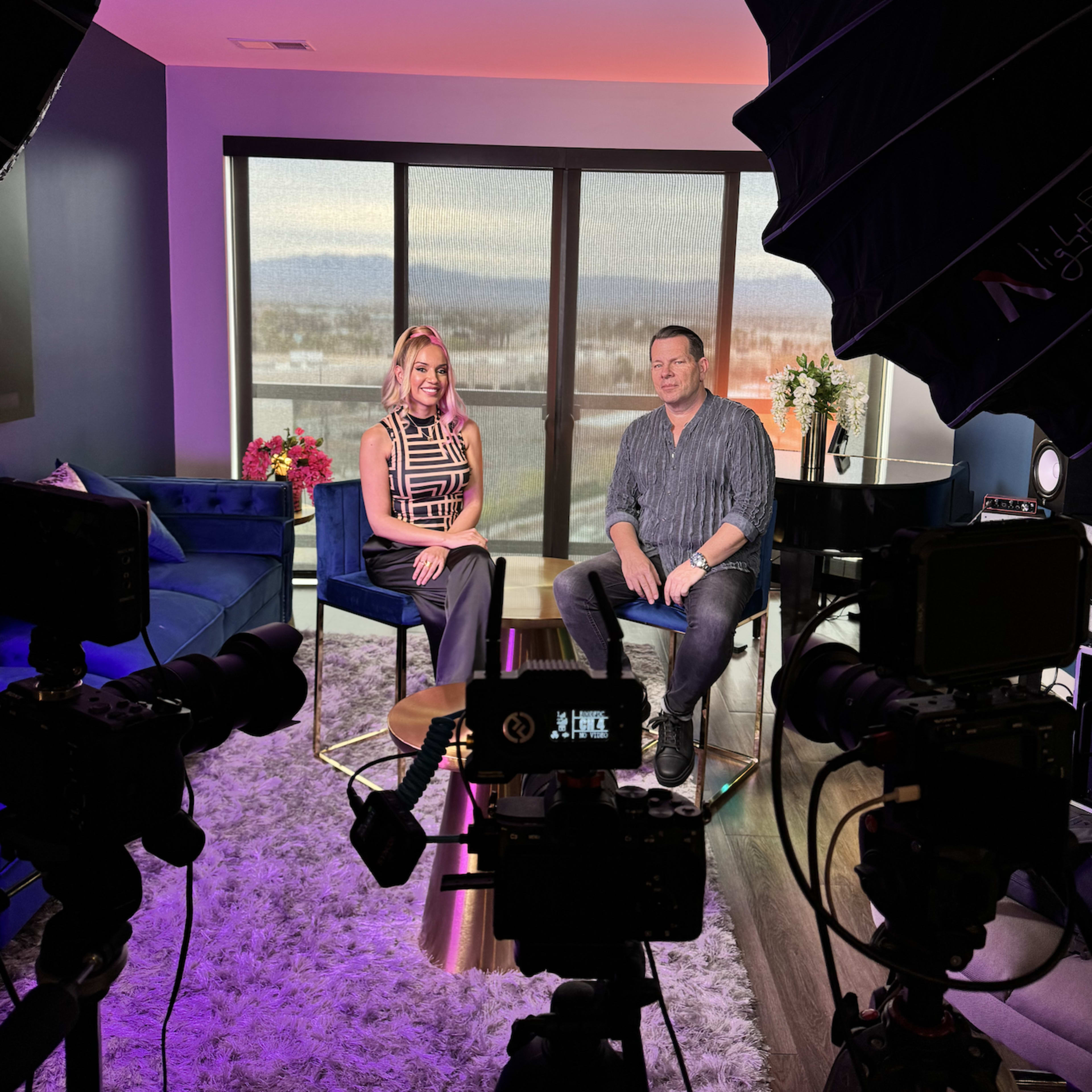 A woman and a man are seated on stools in a well-lit studio with cameras pointed at them and a window displaying a view outside.