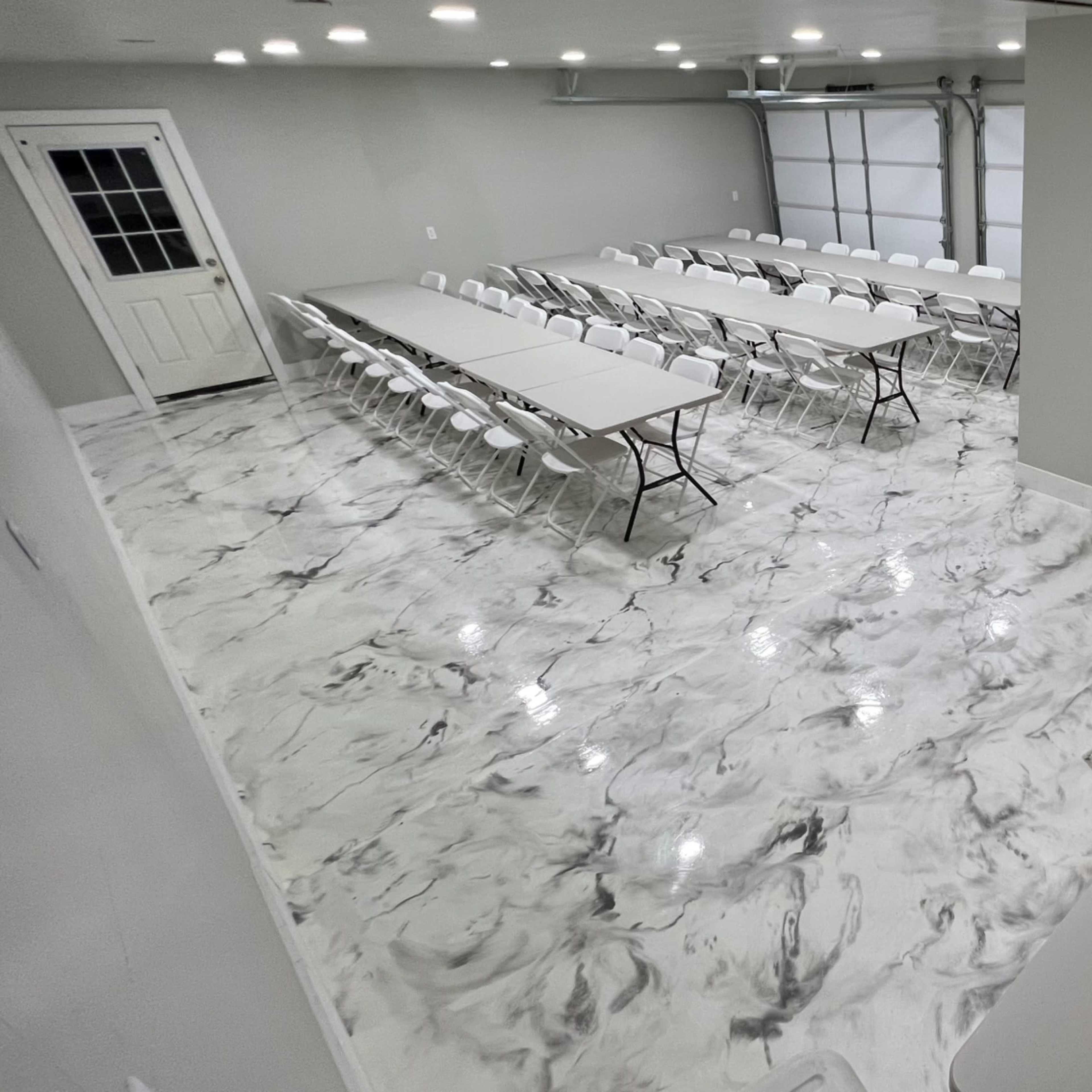 A spacious room with a marble-patterned floor, rows of tables and chairs arranged for an event, and a decorative balloon arrangement in one corner.