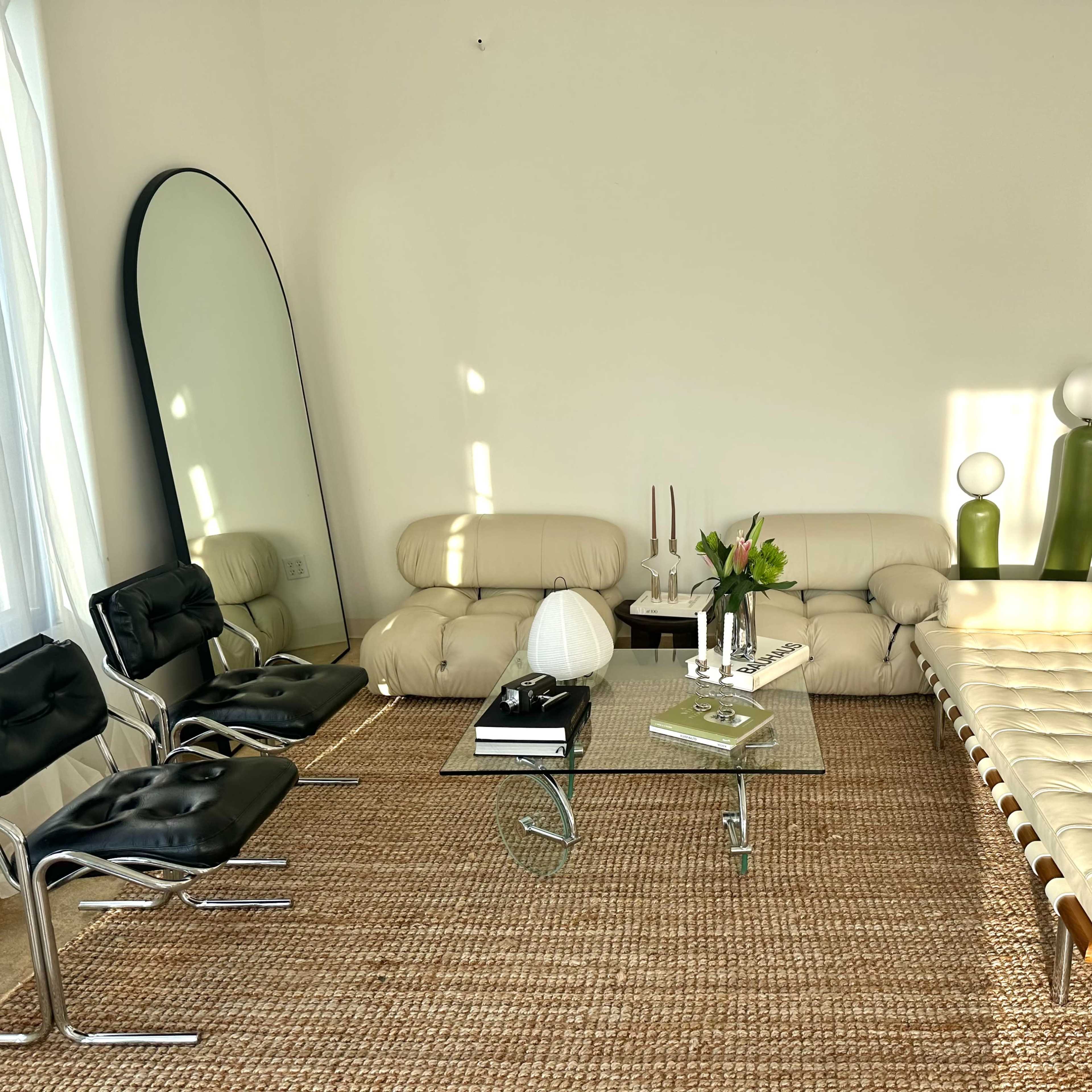 The room features a modern seating area with a cream-colored sofa, black chairs, a glass coffee table, and decorative items, all illuminated by natural light.