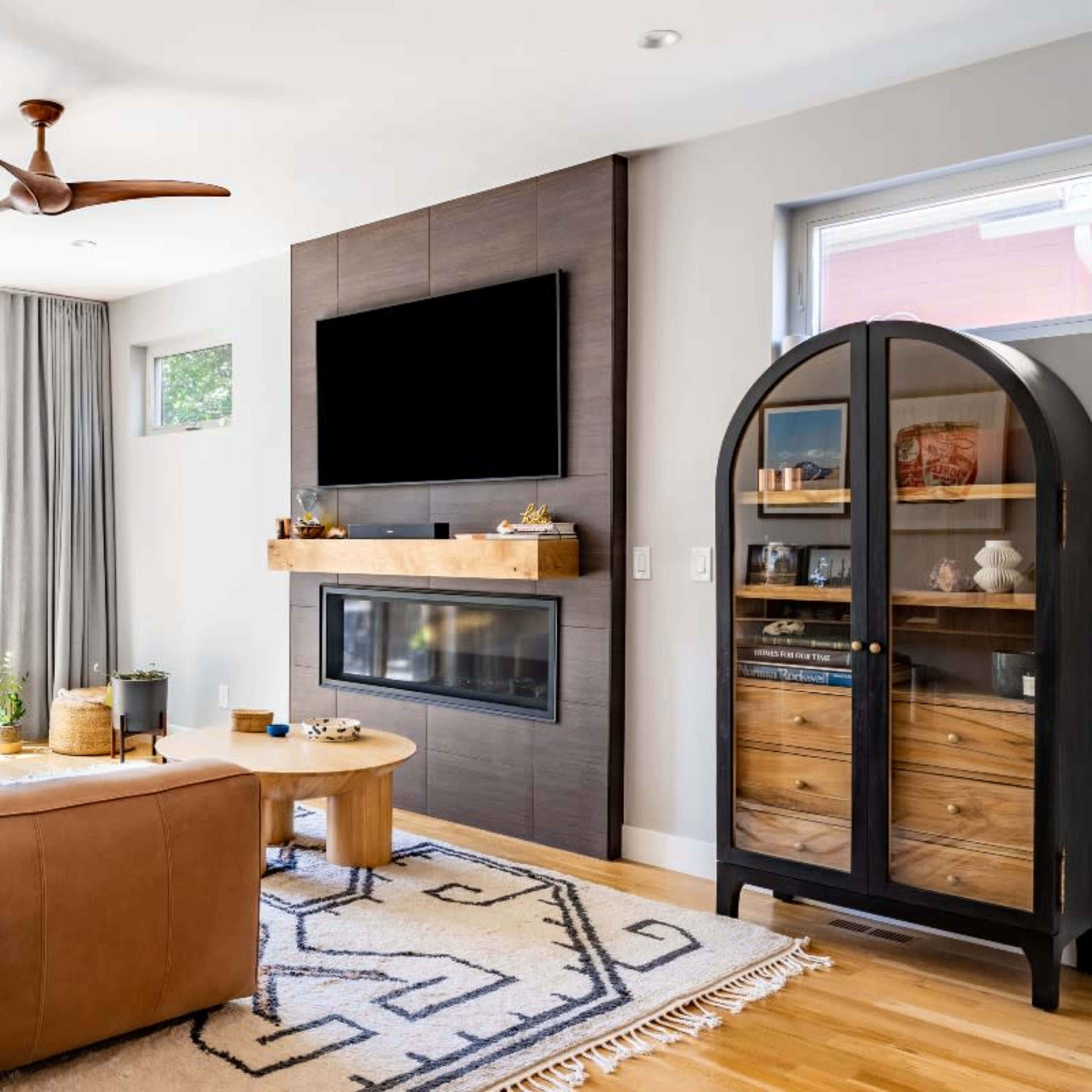 A modern living room features a brown leather sofa, a wall-mounted television above a sleek fireplace, and a glass-front cabinet displaying decorative items, with sliding doors leading to an outdoor space.