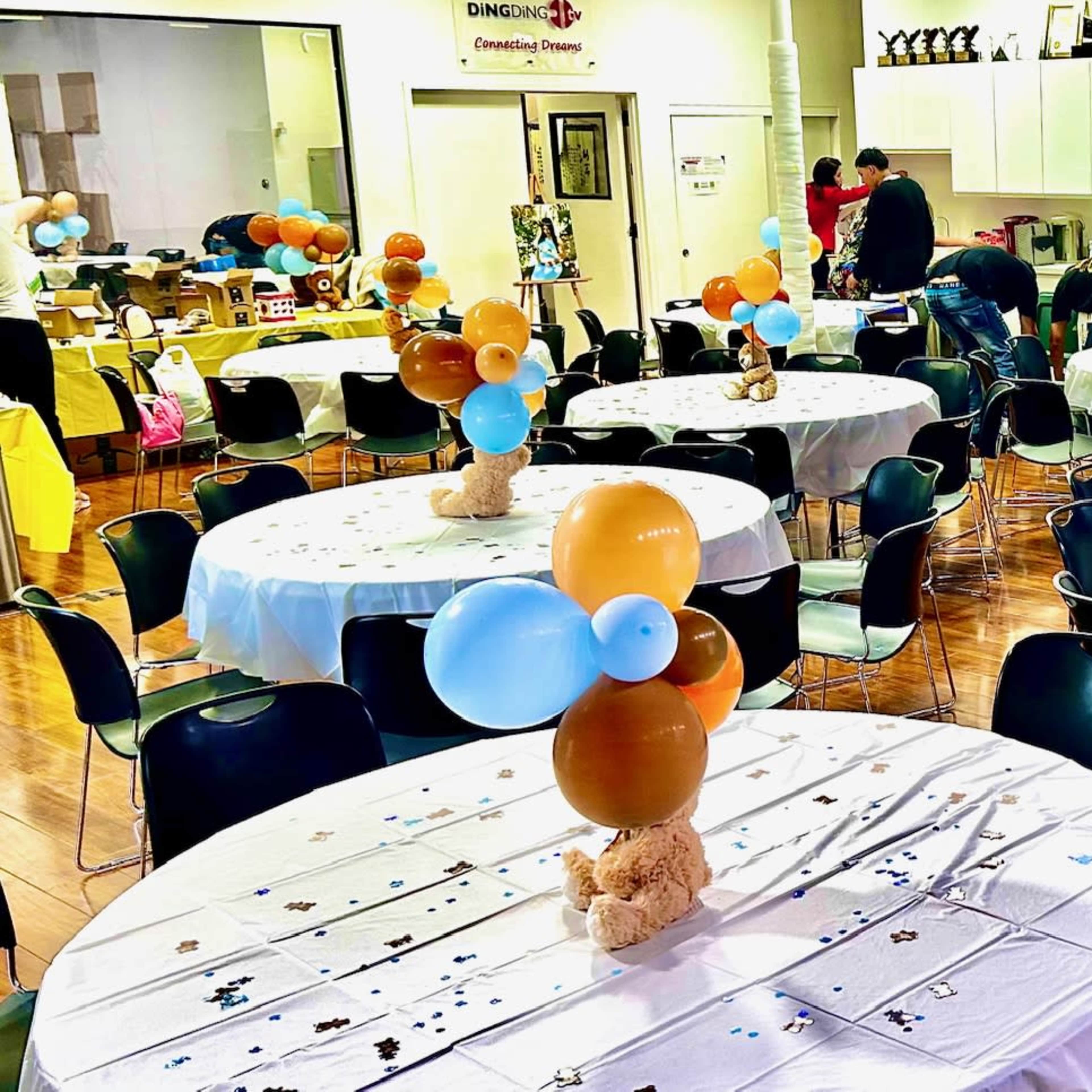 A banquet hall with several round tables covered in white tablecloths, each decorated with clusters of balloons and a small teddy bear centerpiece.