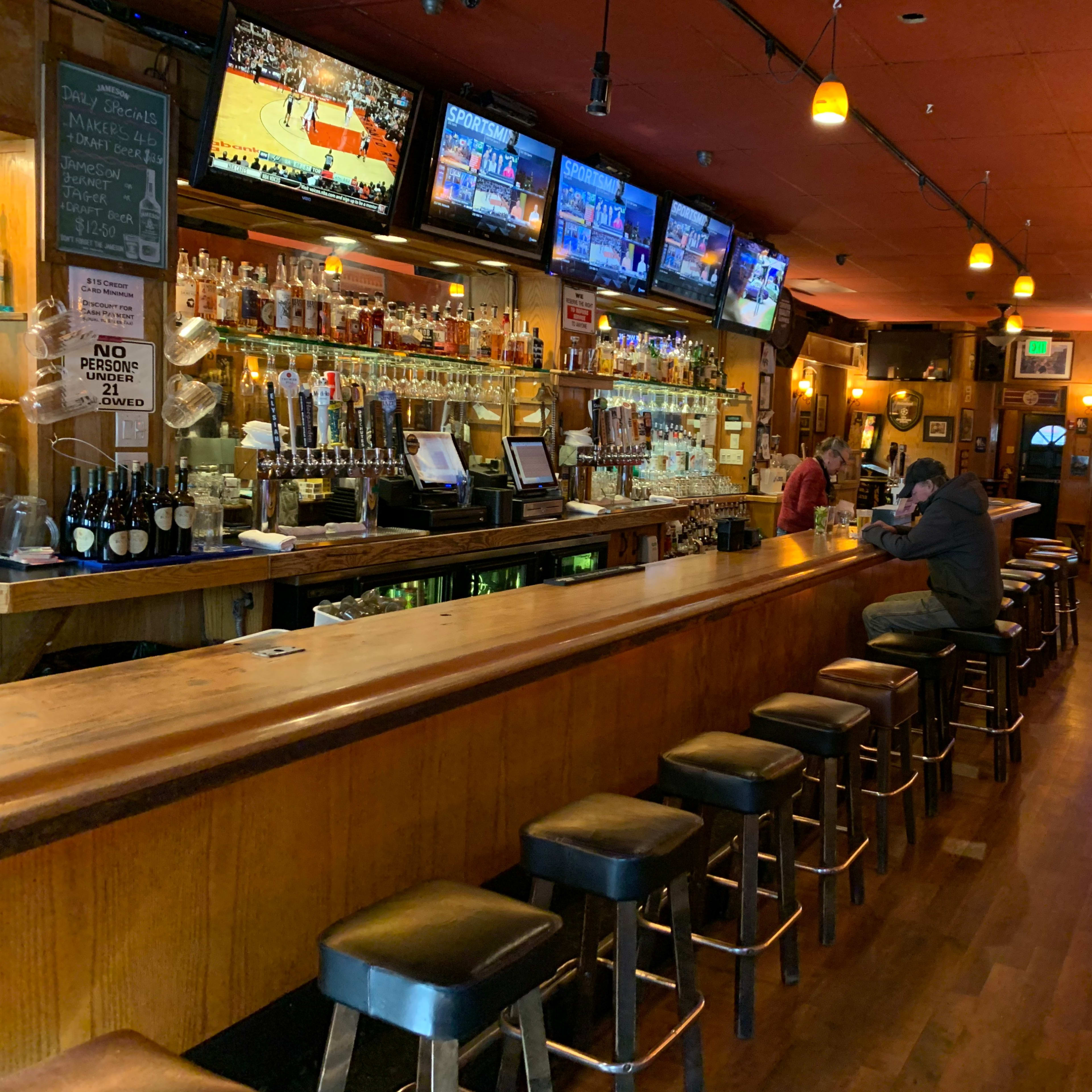 A bar with a long wooden countertop, several high stools, and multiple television screens displaying sports events.