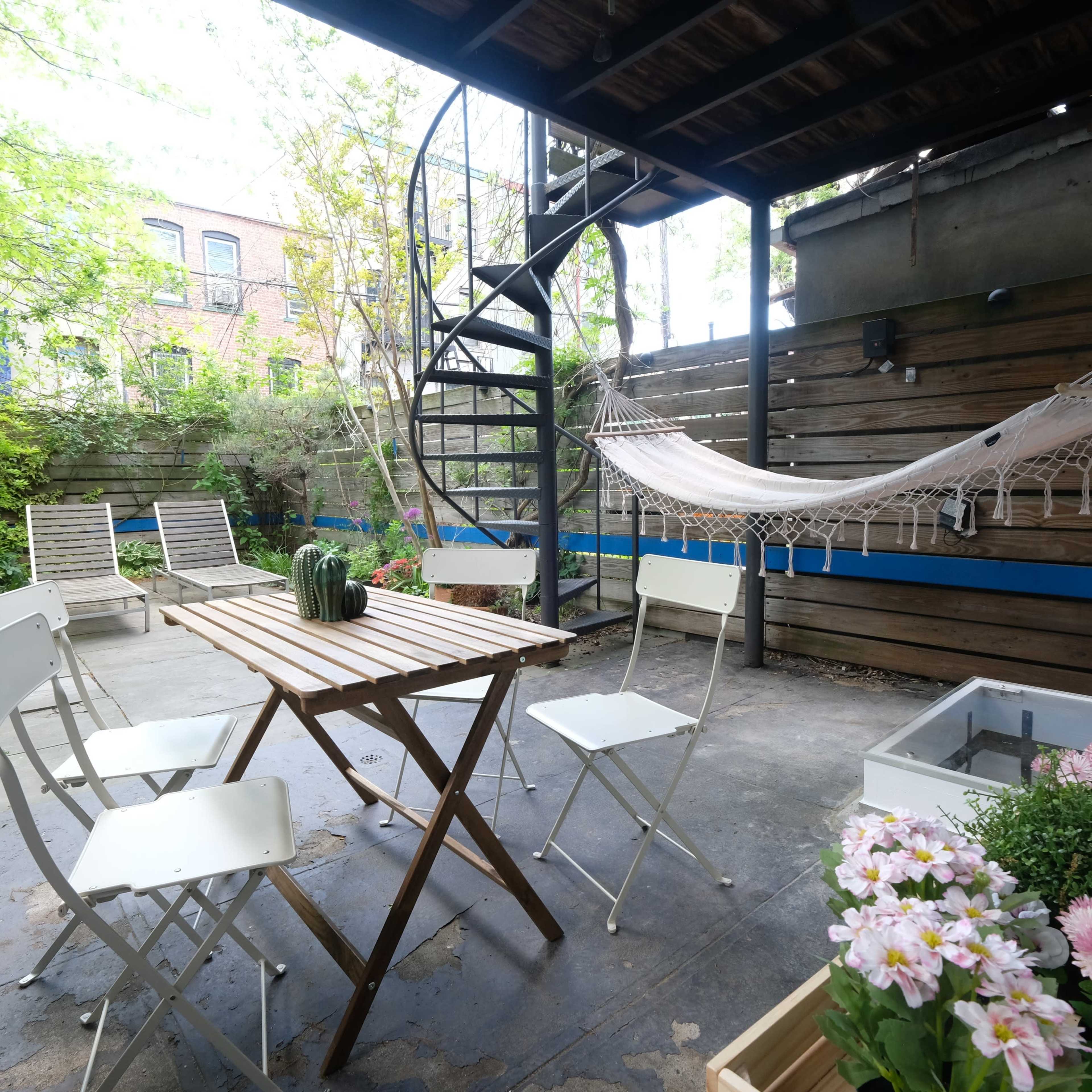 A small outdoor patio area with a spiral staircase, a wooden table surrounded by white chairs, a hammock, and potted flowers.
