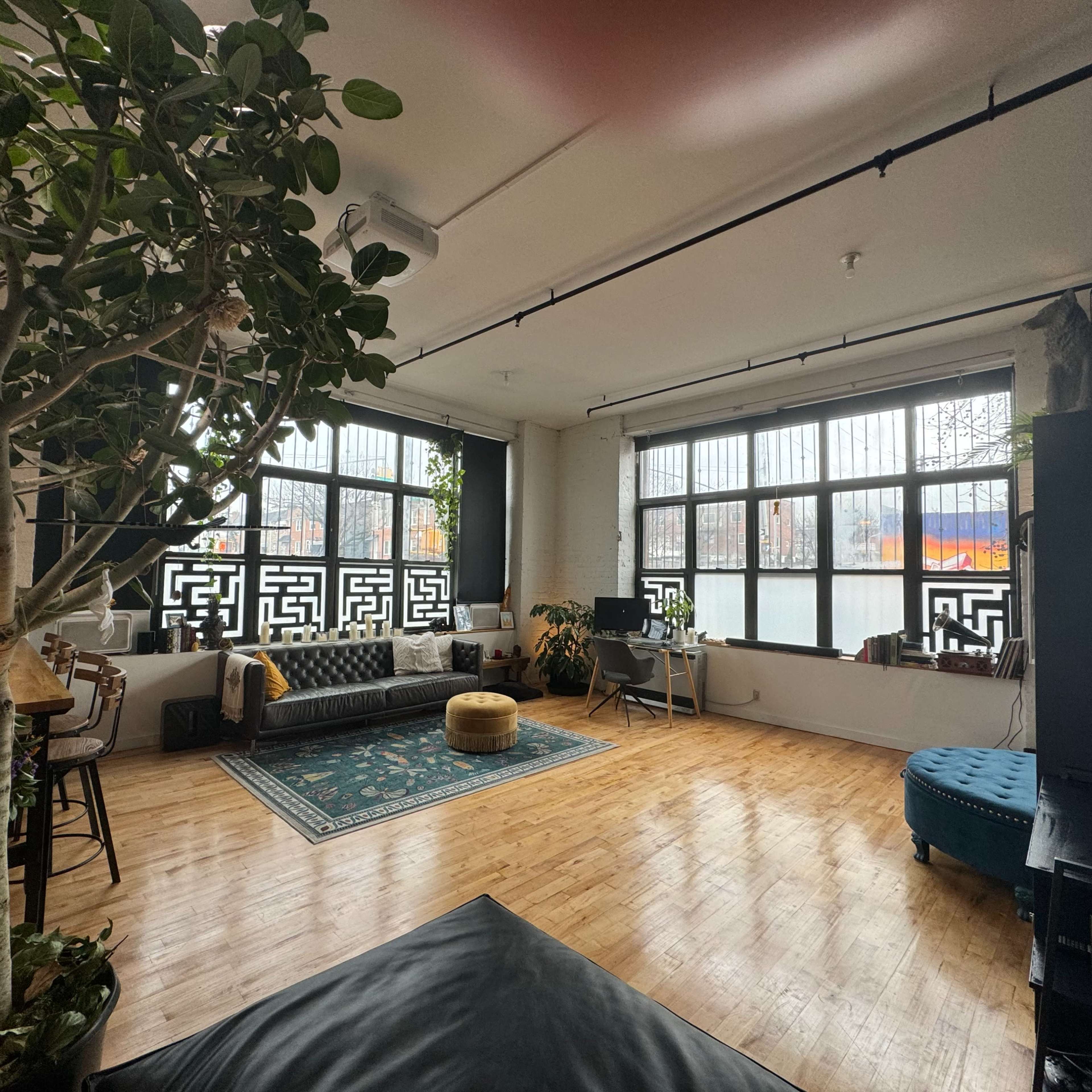 A spacious interior with large windows, a dark sofa, a blue ottoman, and plants, along with wooden flooring.