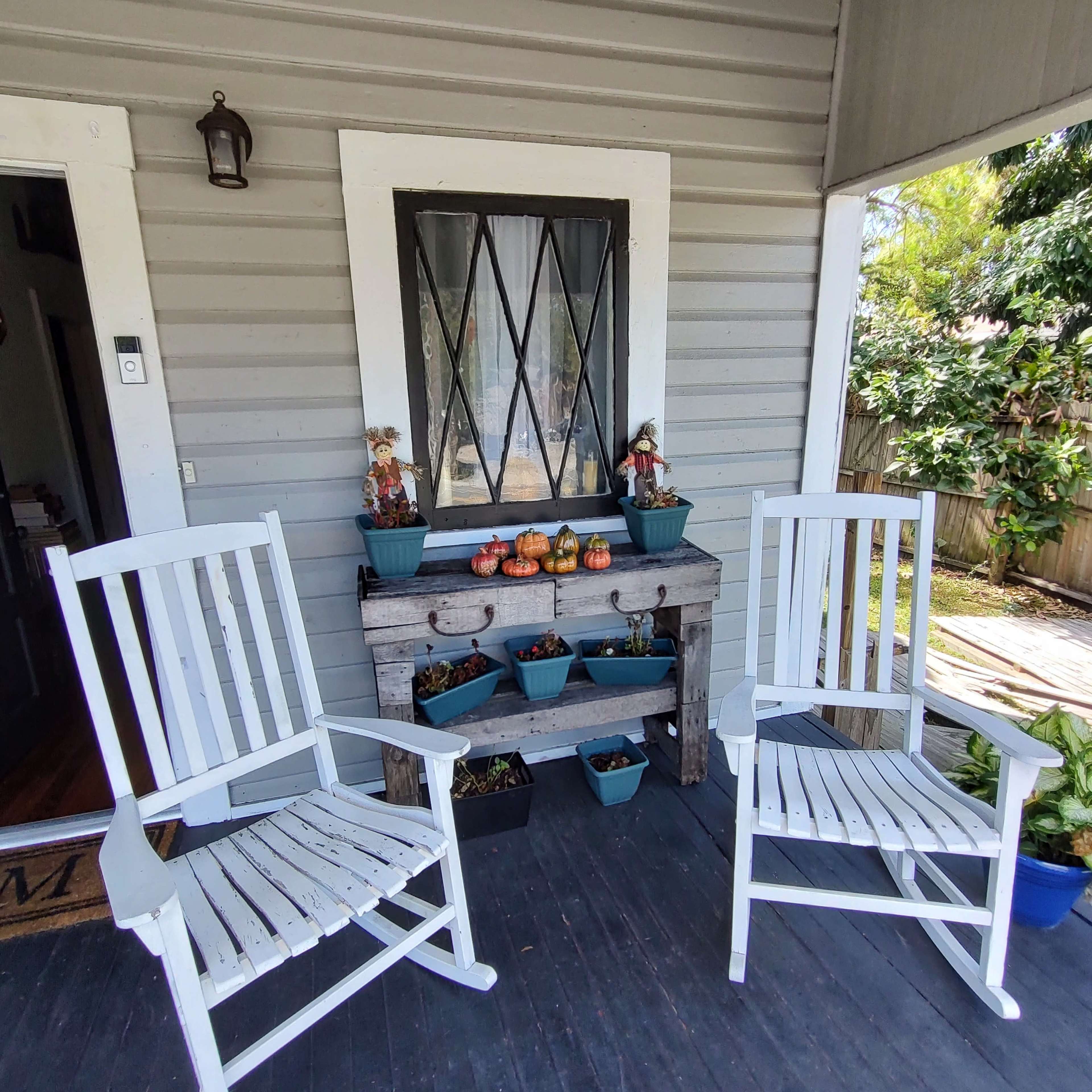 Two white rocking chairs sit on a wooden porch in front of a table adorned with potted plants and decorative items.