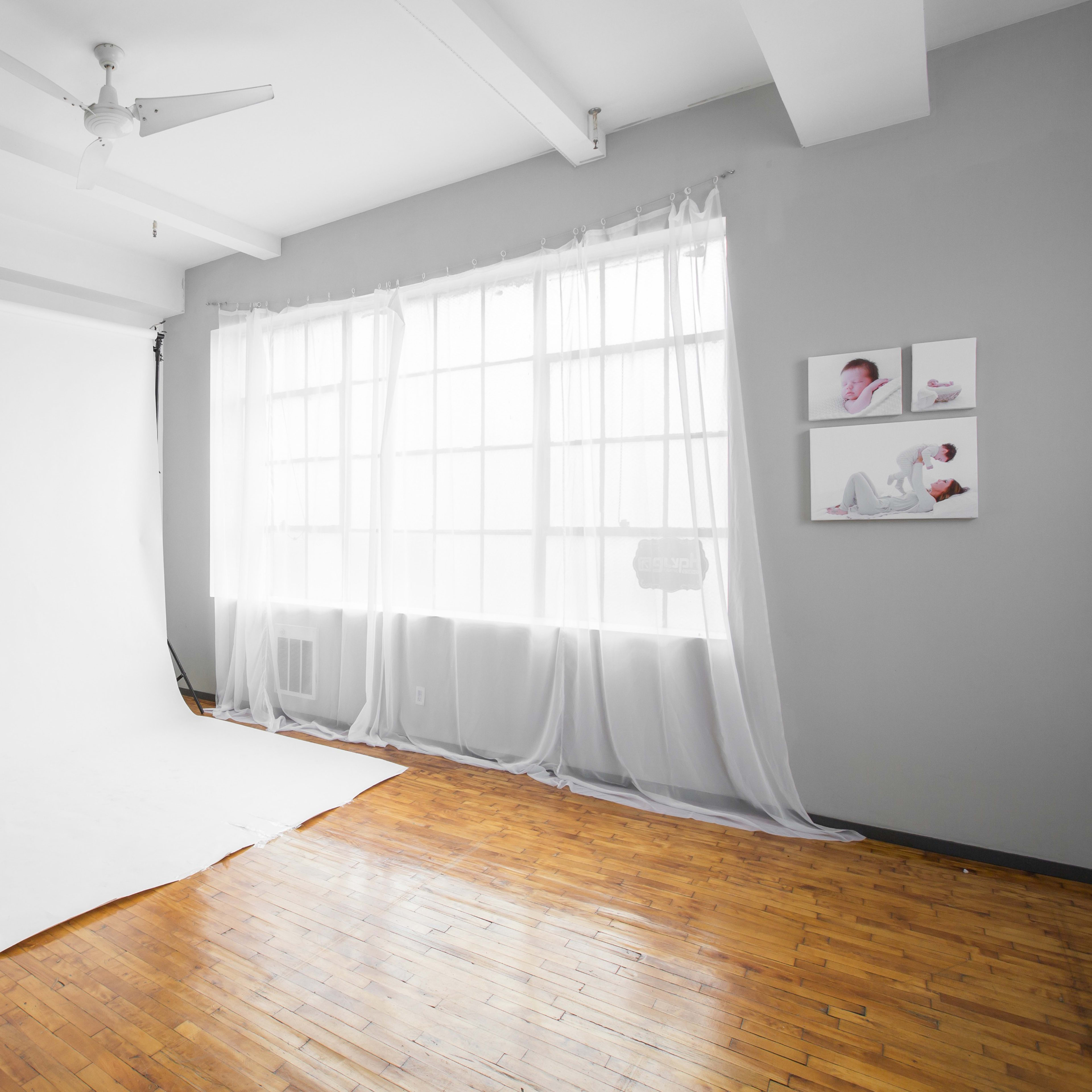 A bright, minimalistic photography studio with a white backdrop, hardwood flooring, and large windows.