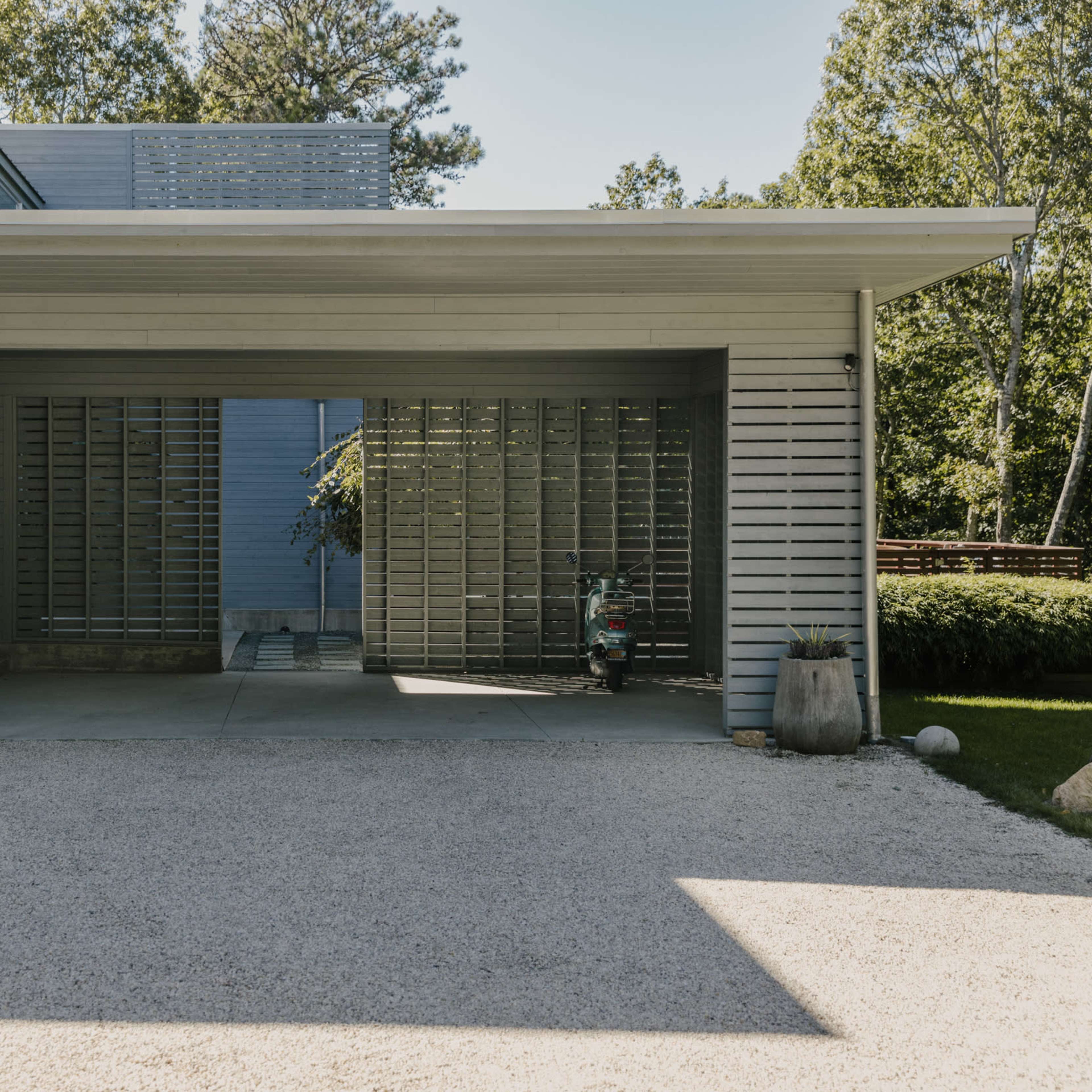 A modern gray wooden structure with a covered parking area and a gravel driveway, surrounded by greenery.