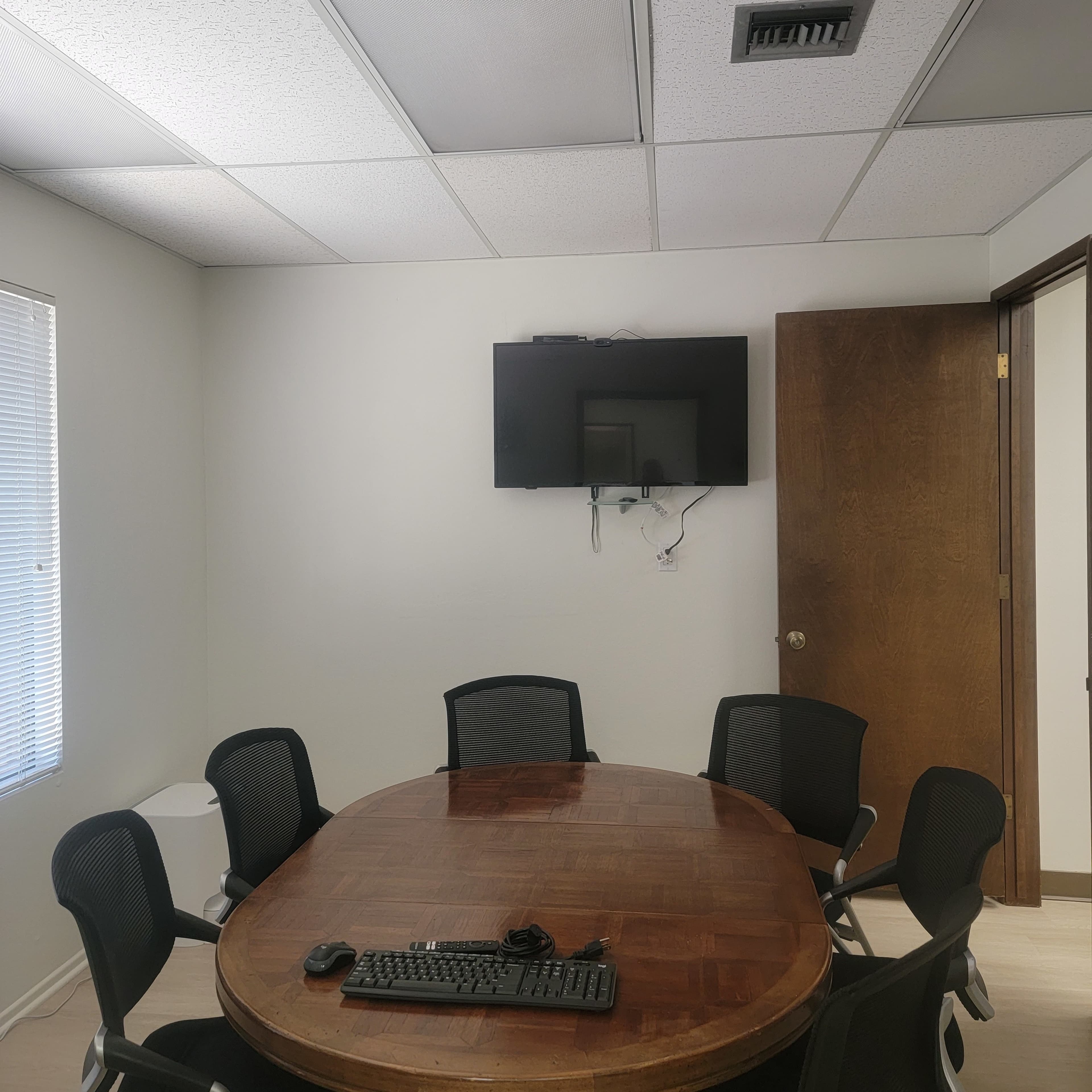 A conference room features a round wooden table surrounded by six black chairs, with a television mounted on the wall.