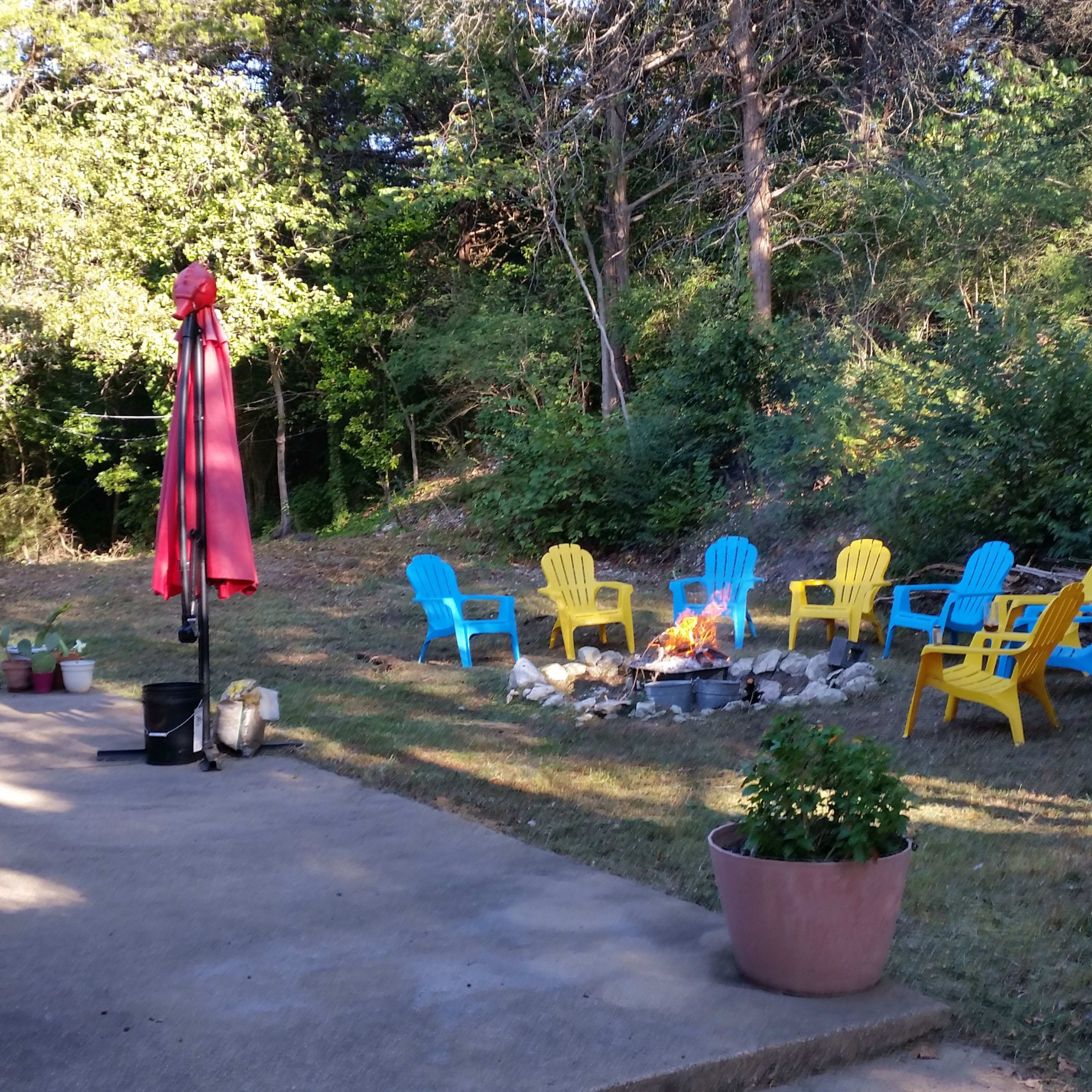 A patio area features a circular fire pit surrounded by vibrant blue and yellow chairs, with a red umbrella nearby and potted plants in the foreground.