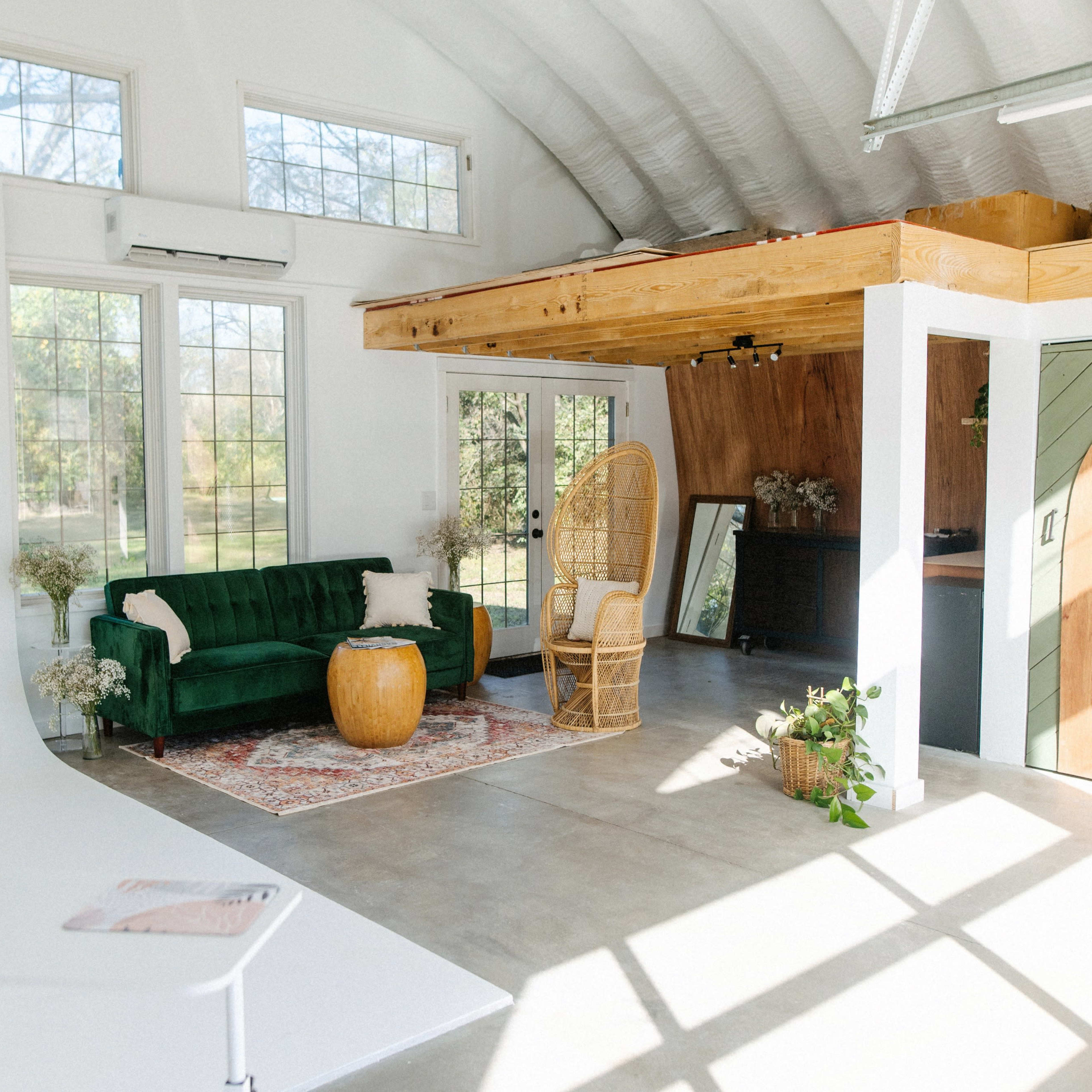 A bright, modern interior featuring a green sofa, a rattan chair, and large windows allowing natural light to fill the space.