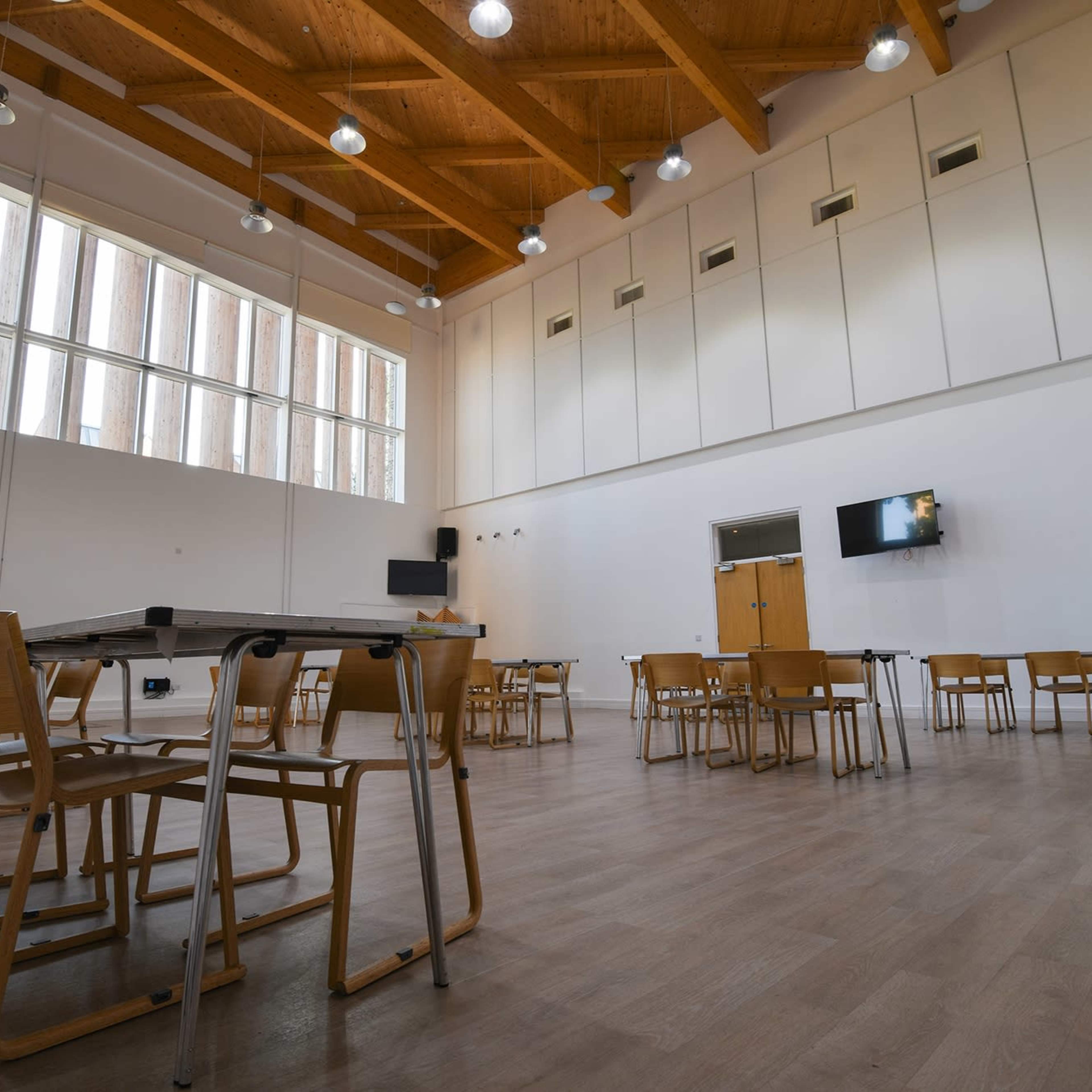 A spacious meeting room features wooden beams, several tables, and chairs arranged on a light-colored floor.