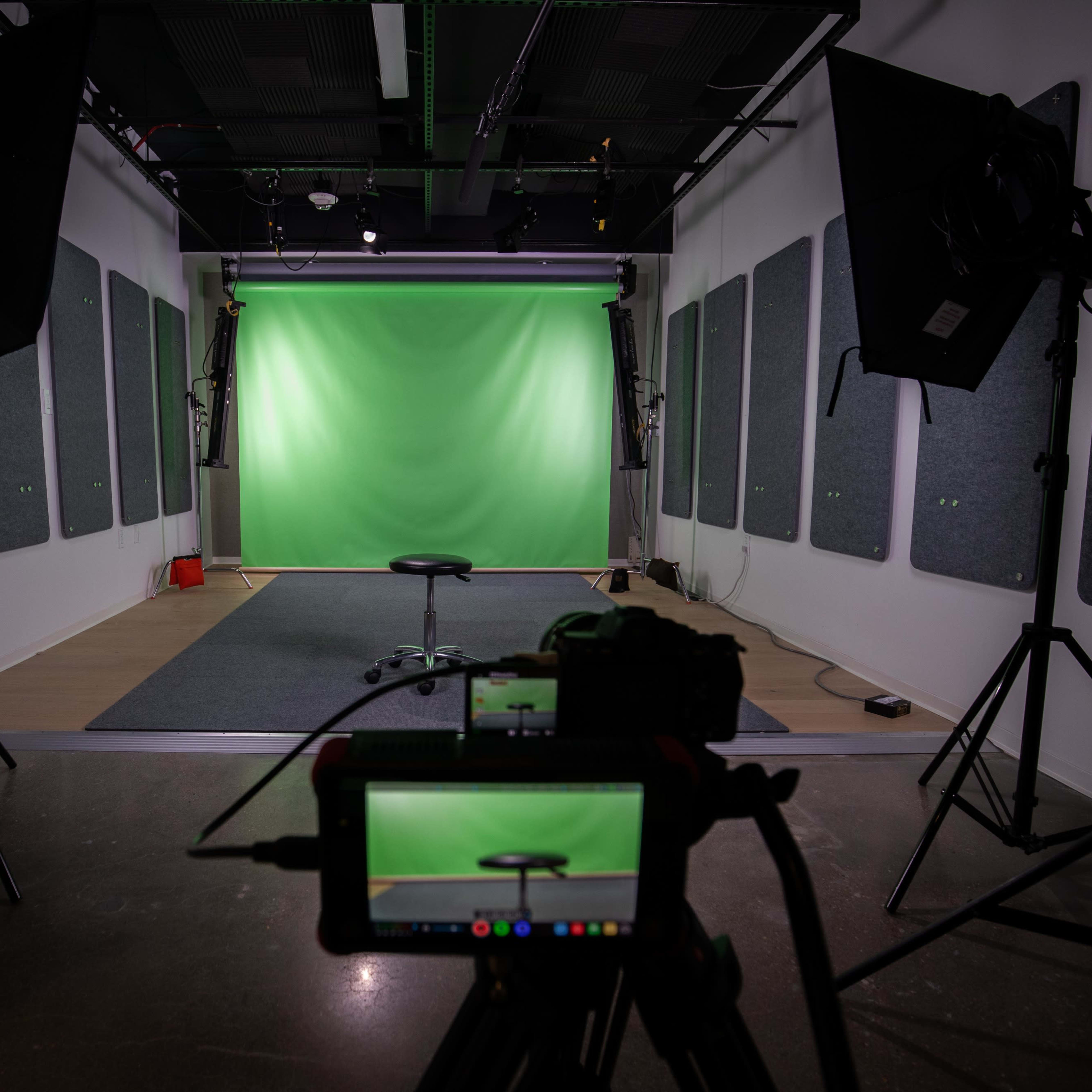 A studio setup with a green screen backdrop, a black stool in the center, and professional lighting equipment on either side.