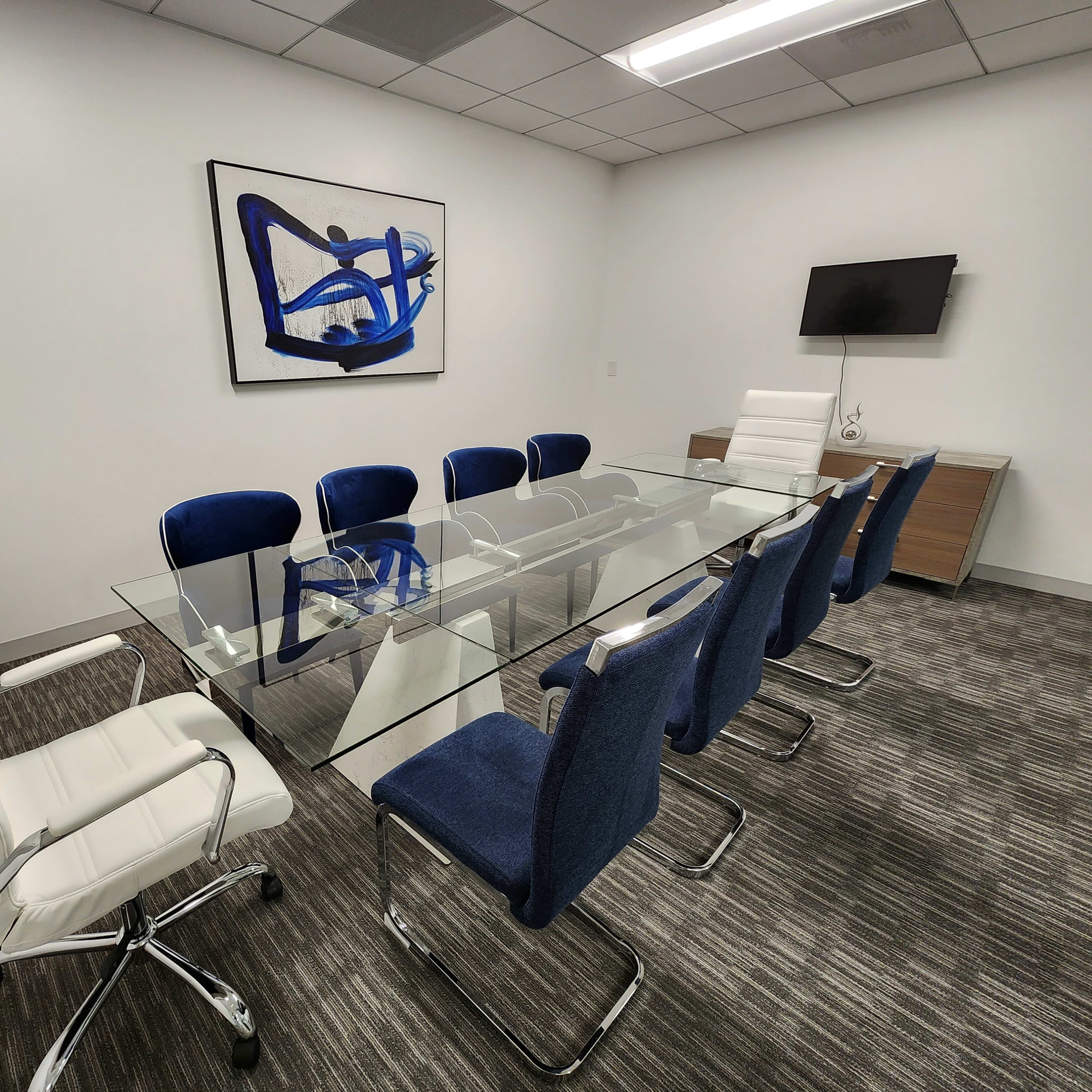 A modern conference room features a glass table surrounded by blue upholstered chairs and a white leather chair, with a large abstract painting on the wall.