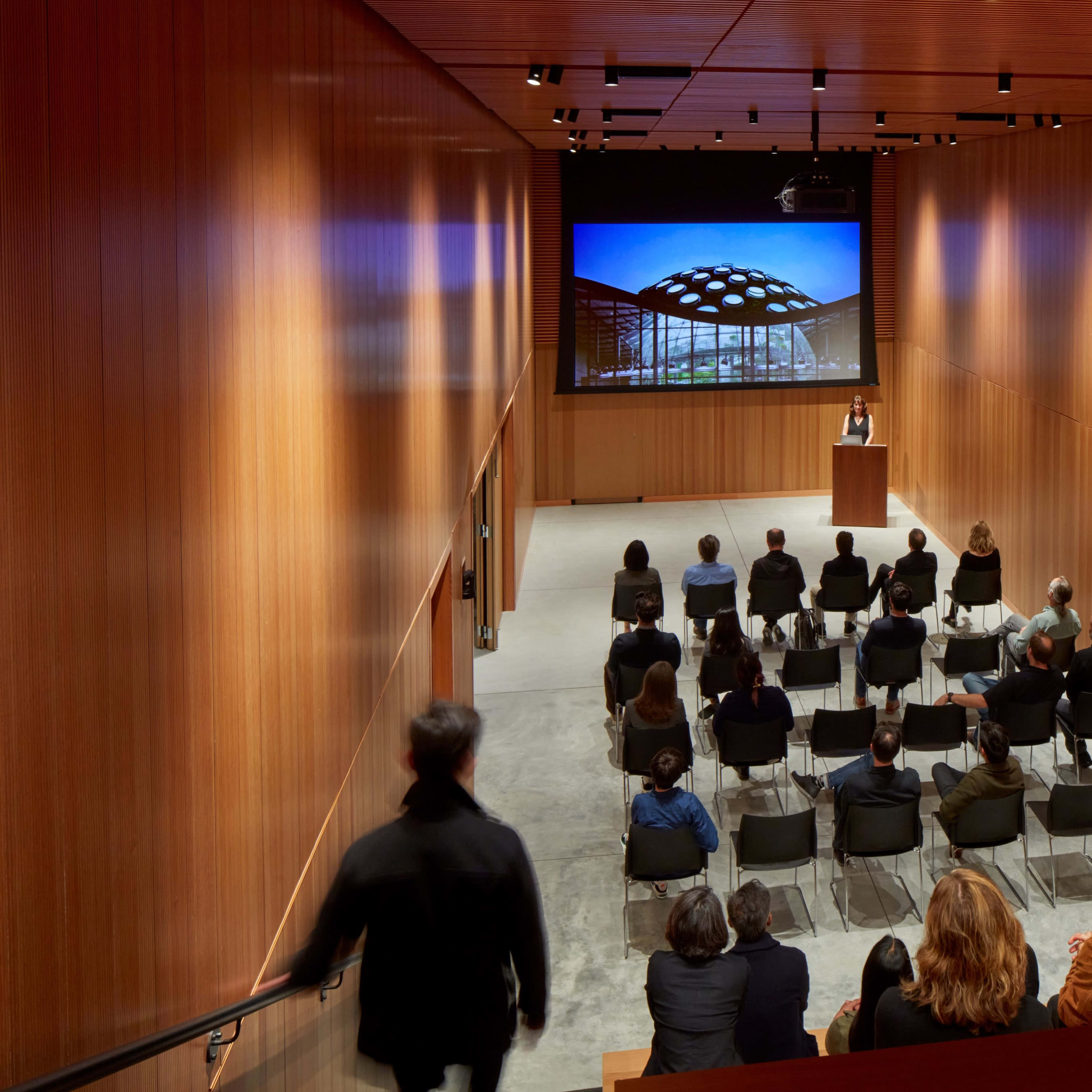 A speaker presents to an audience in a modern auditorium with wooden walls and a large screen displaying images.