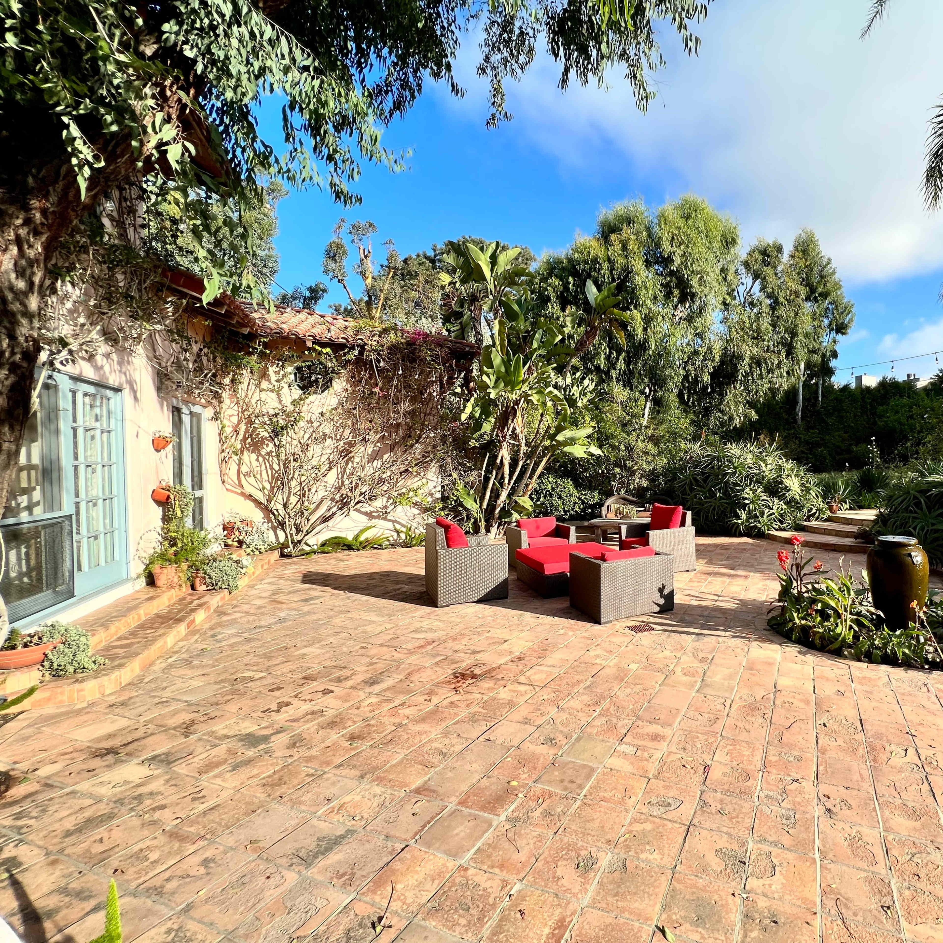 A patio with red chairs and a table is surrounded by various plants and trees under a partly cloudy sky.