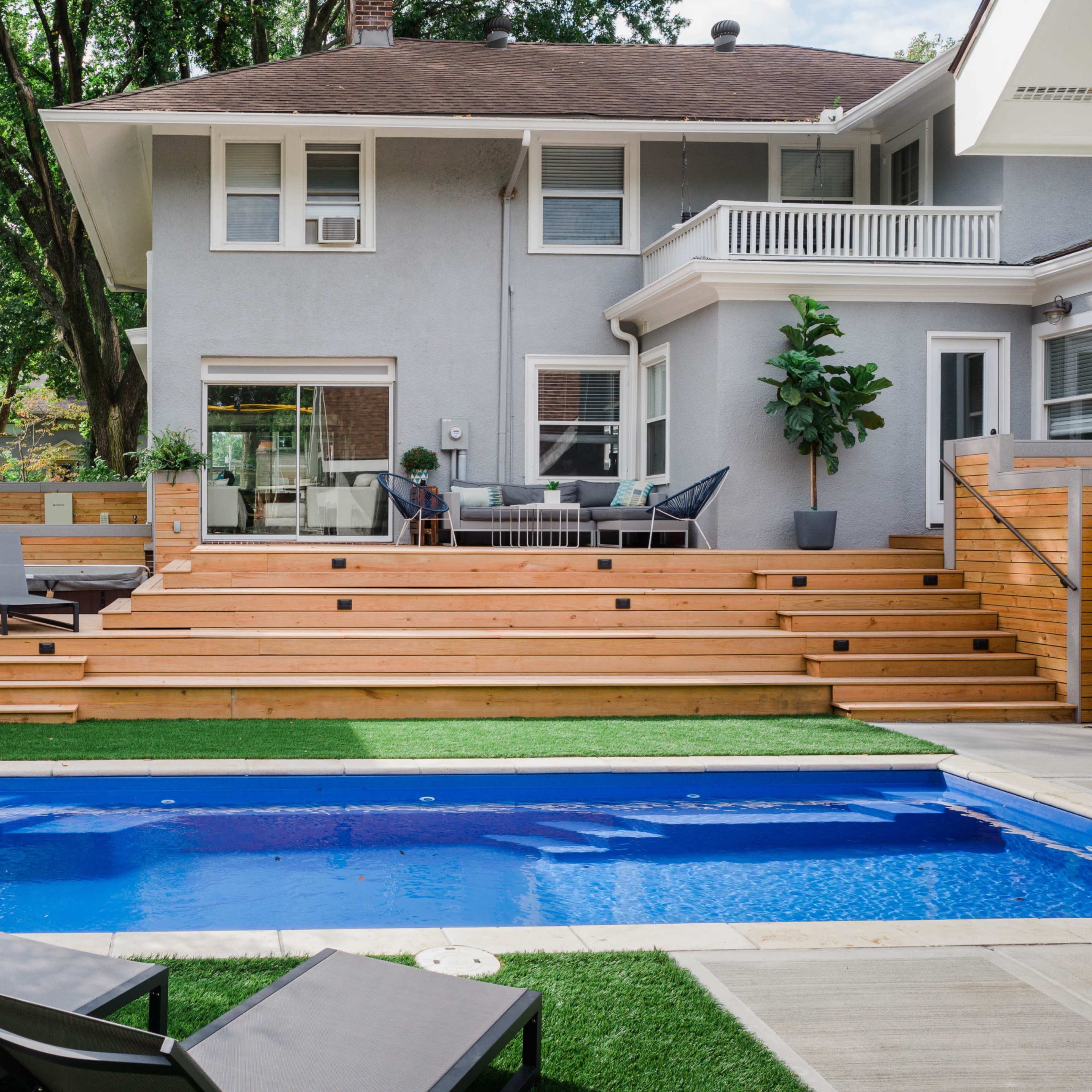 A backyard features a wooden deck leading to a rectangular swimming pool, surrounded by grass and several lounge chairs.