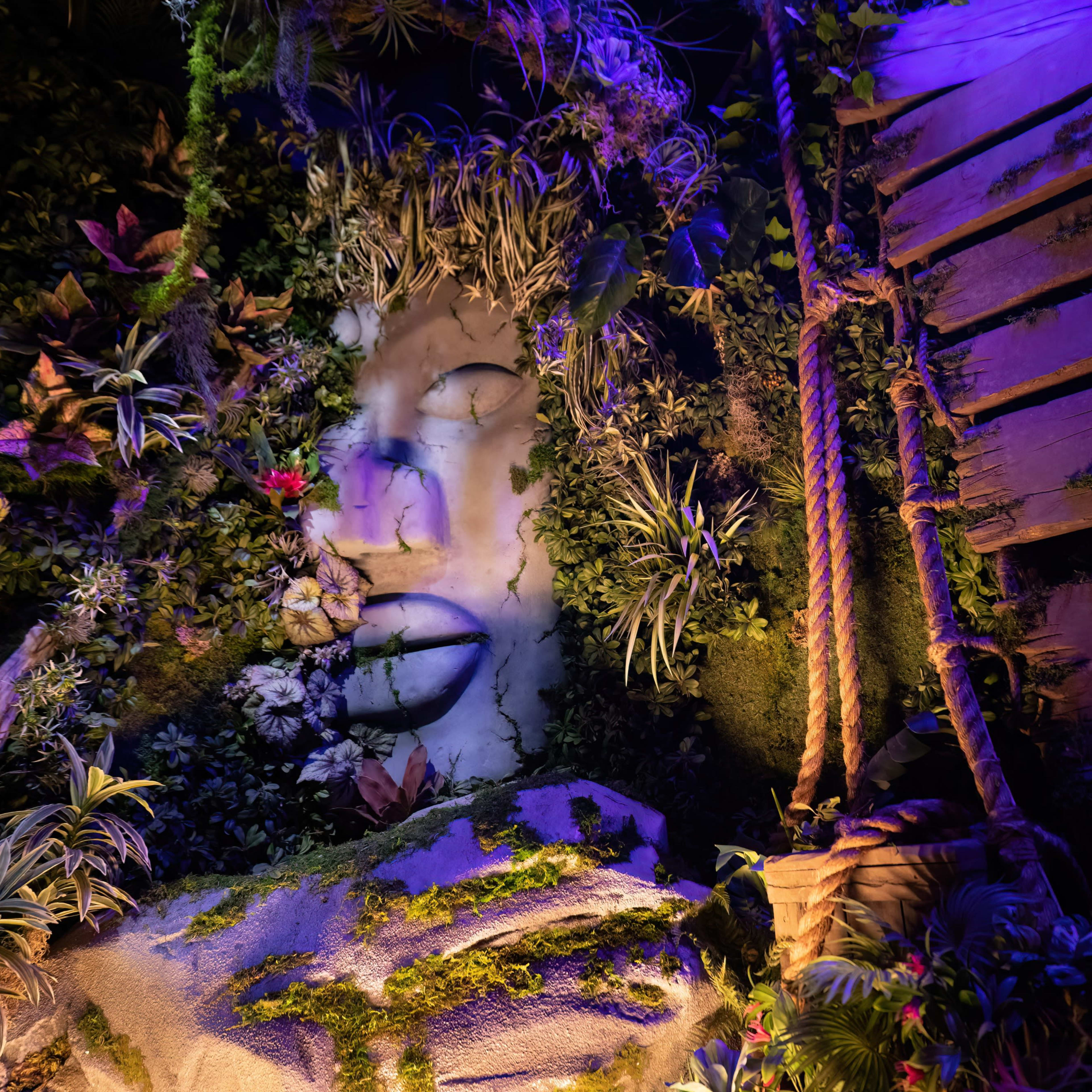 A large, artistic face carved into a rock surface is surrounded by vibrant foliage and illuminated by colored lights, with a wooden structure to the side.