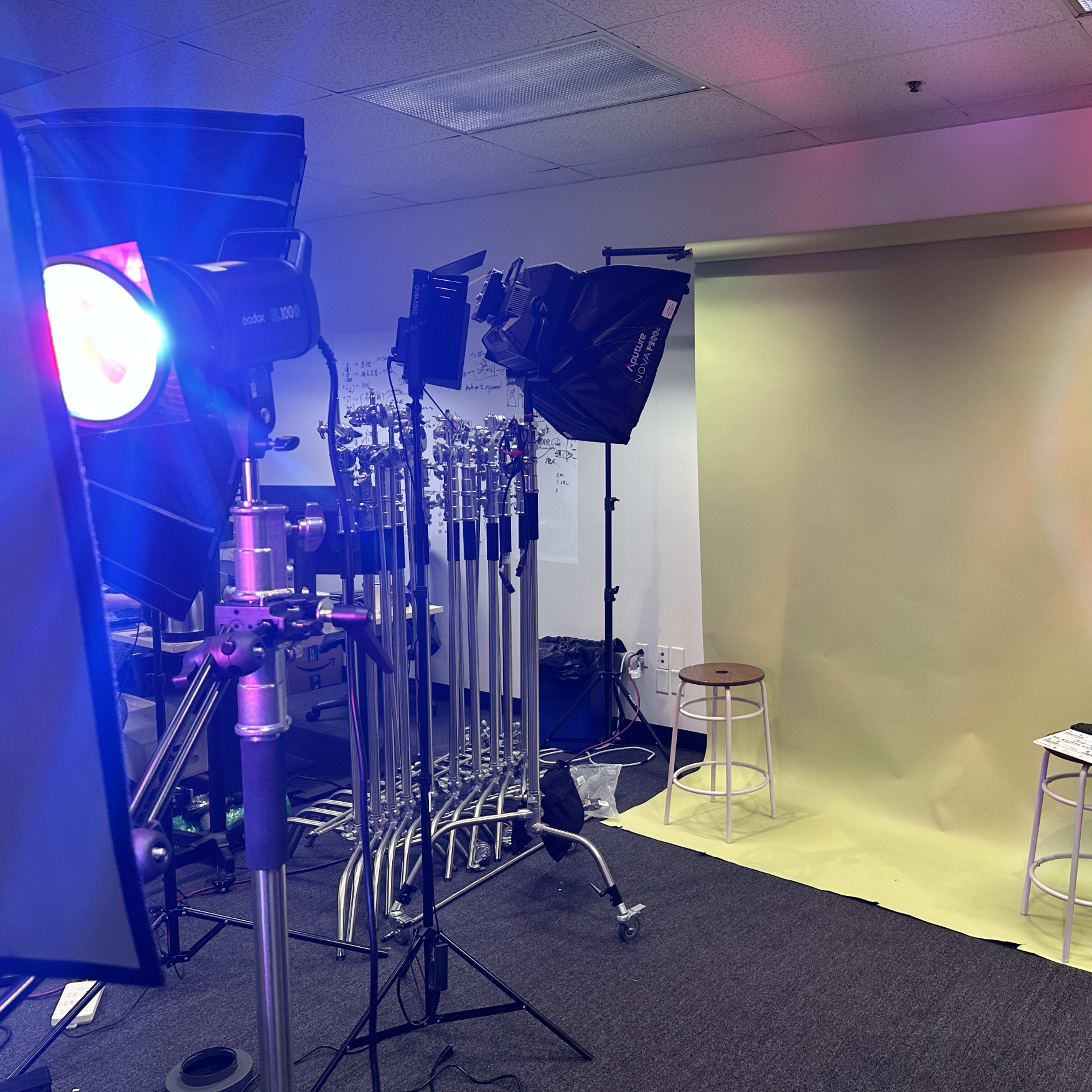 A photo studio setup with two stools, a yellow backdrop, and various lighting equipment arranged around the space.