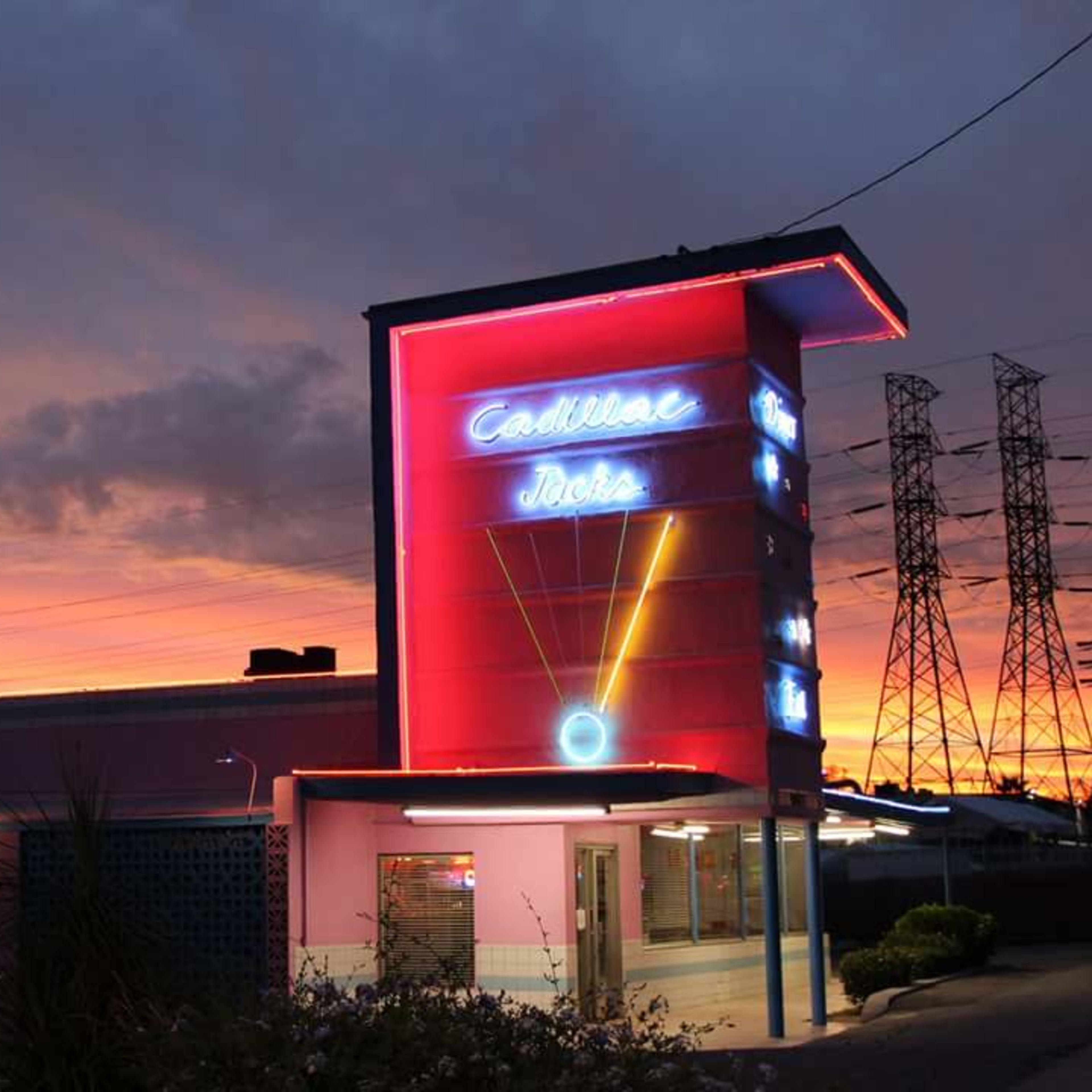 A neon-lit building with a "Cadillac Jack's" sign stands against a colorful sunset sky, surrounded by power lines and trees.
