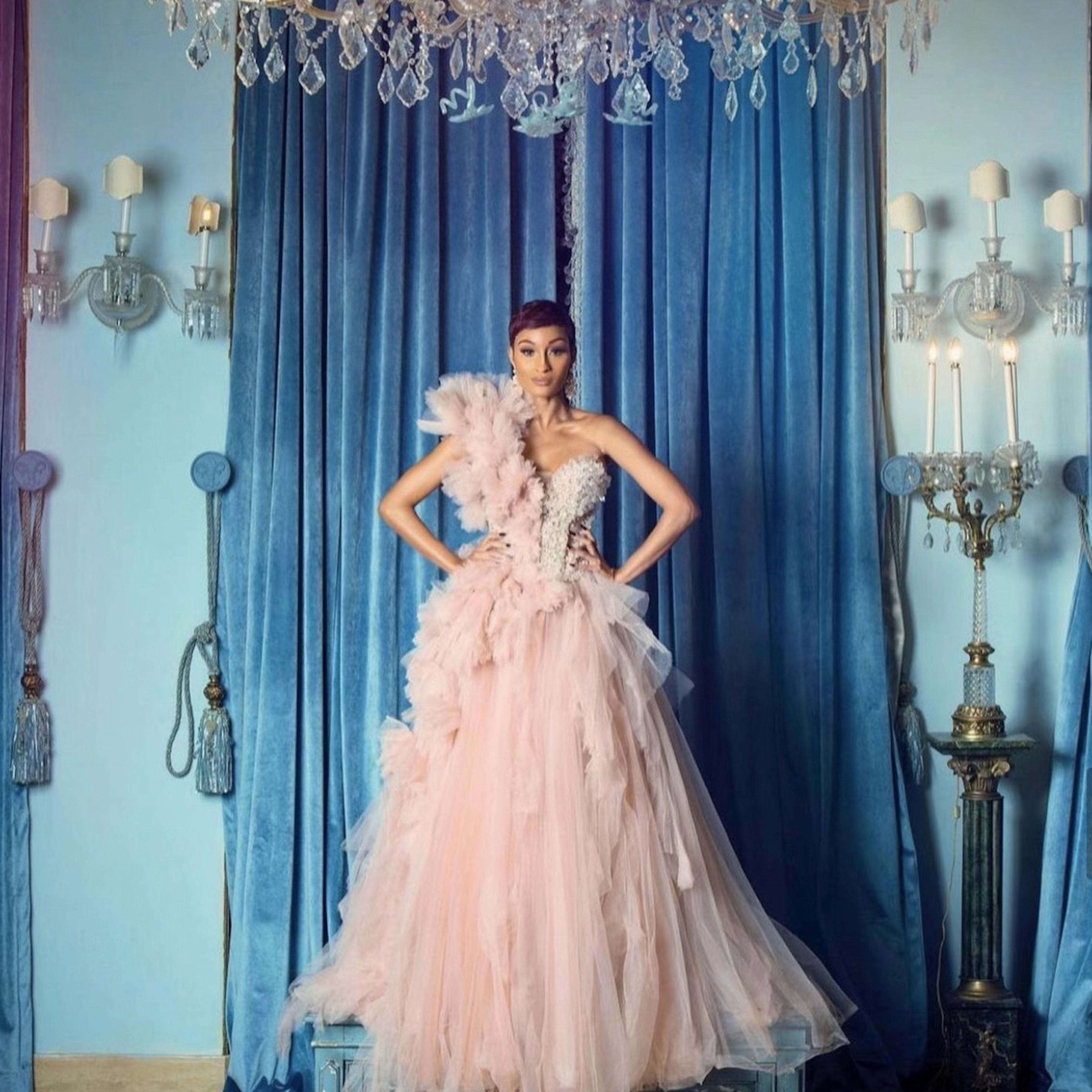 A model poses in an elaborate pink gown with feathers in front of a blue backdrop and ornate decor, including a chandelier and wall sconces.