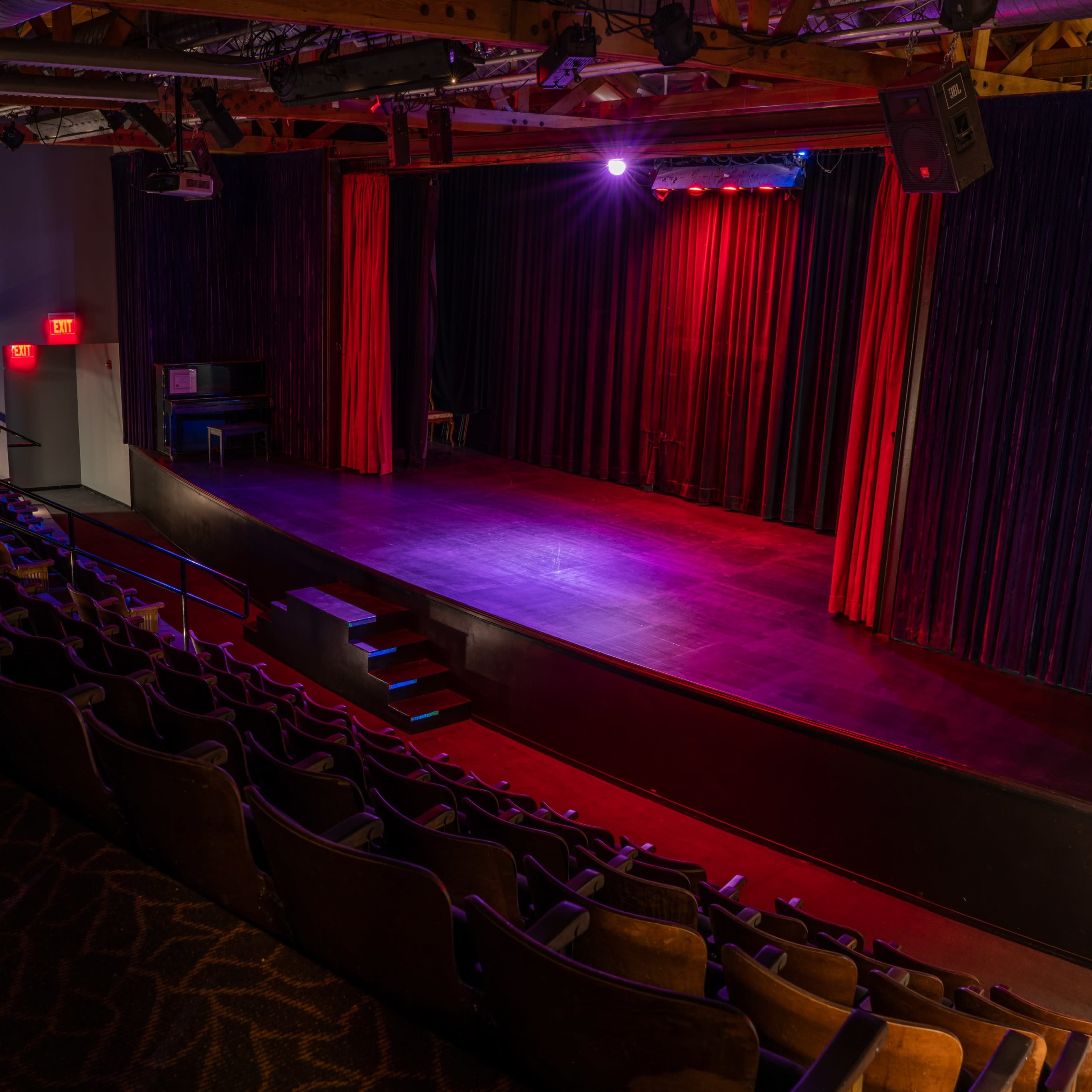 An empty theater with rows of seats facing a small stage illuminated by red and purple lights.