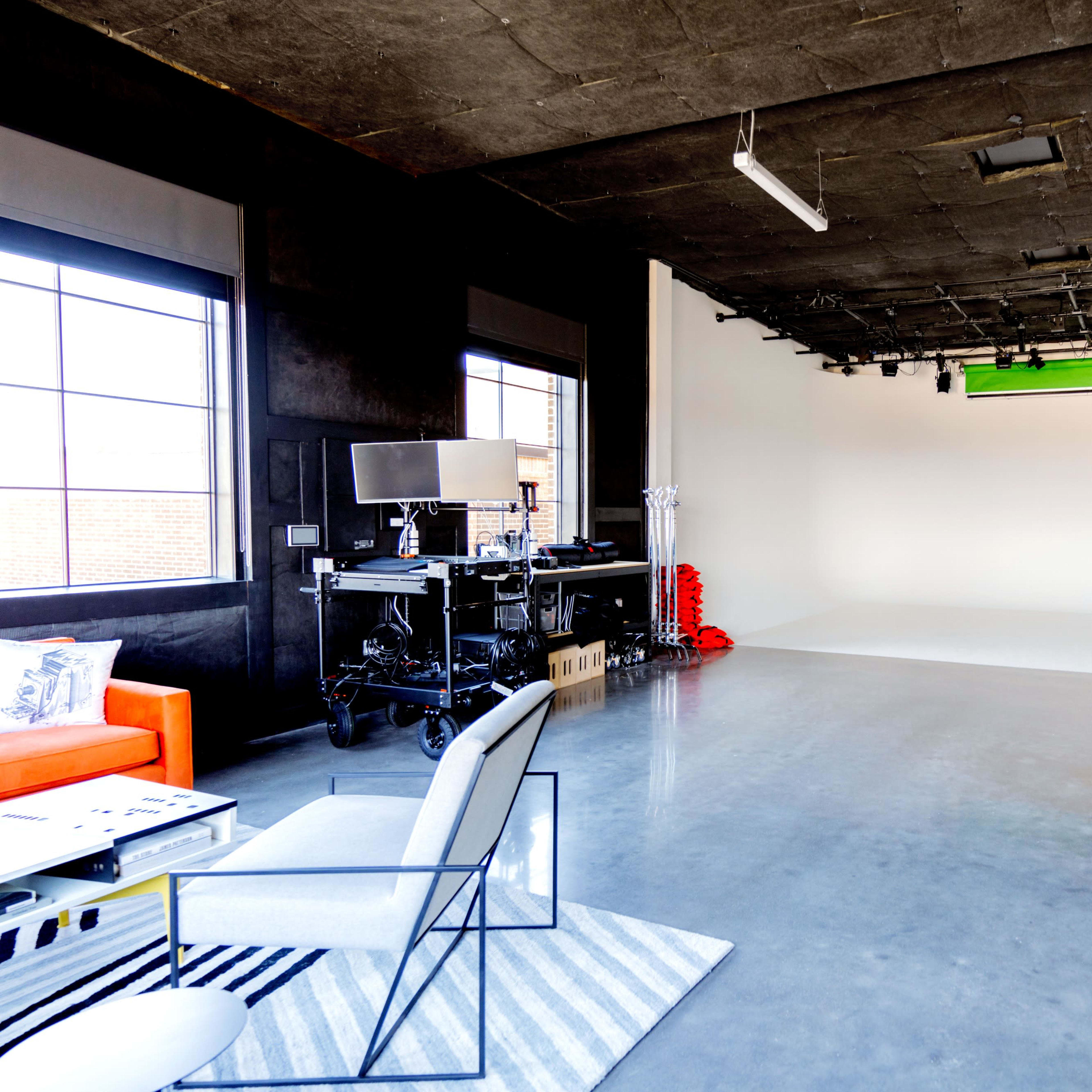 A spacious studio with a concrete floor, a large empty backdrop, and equipment mounted along the walls, including a green screen and lighting setups.