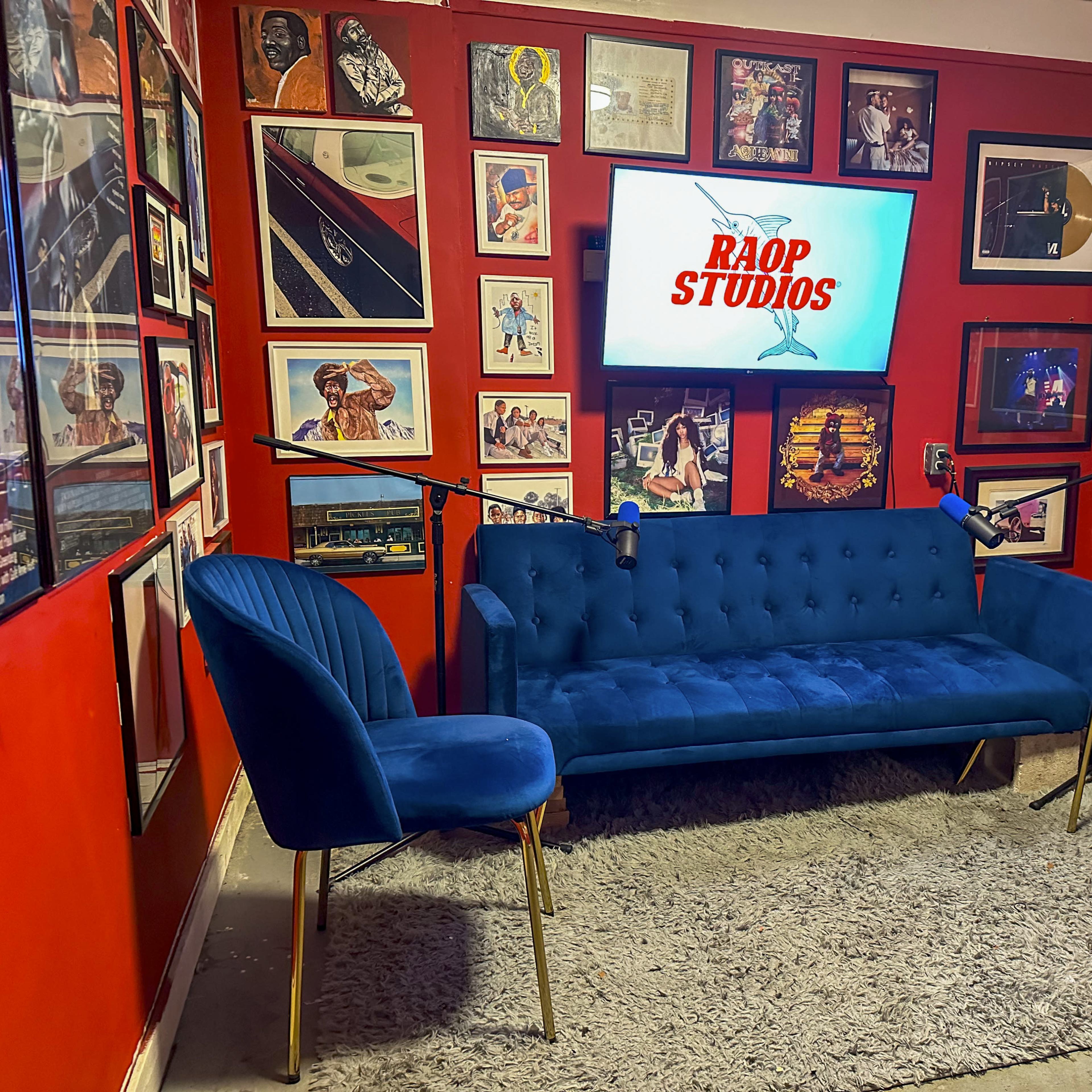 A small recording studio with a red wall adorned with framed posters, a blue sofa, two blue chairs, and a screen displaying "RAOP STUDIOS."